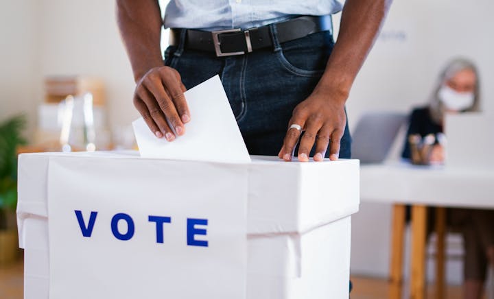 A person is casting a vote into a ballot box with "VOTE" written on it.