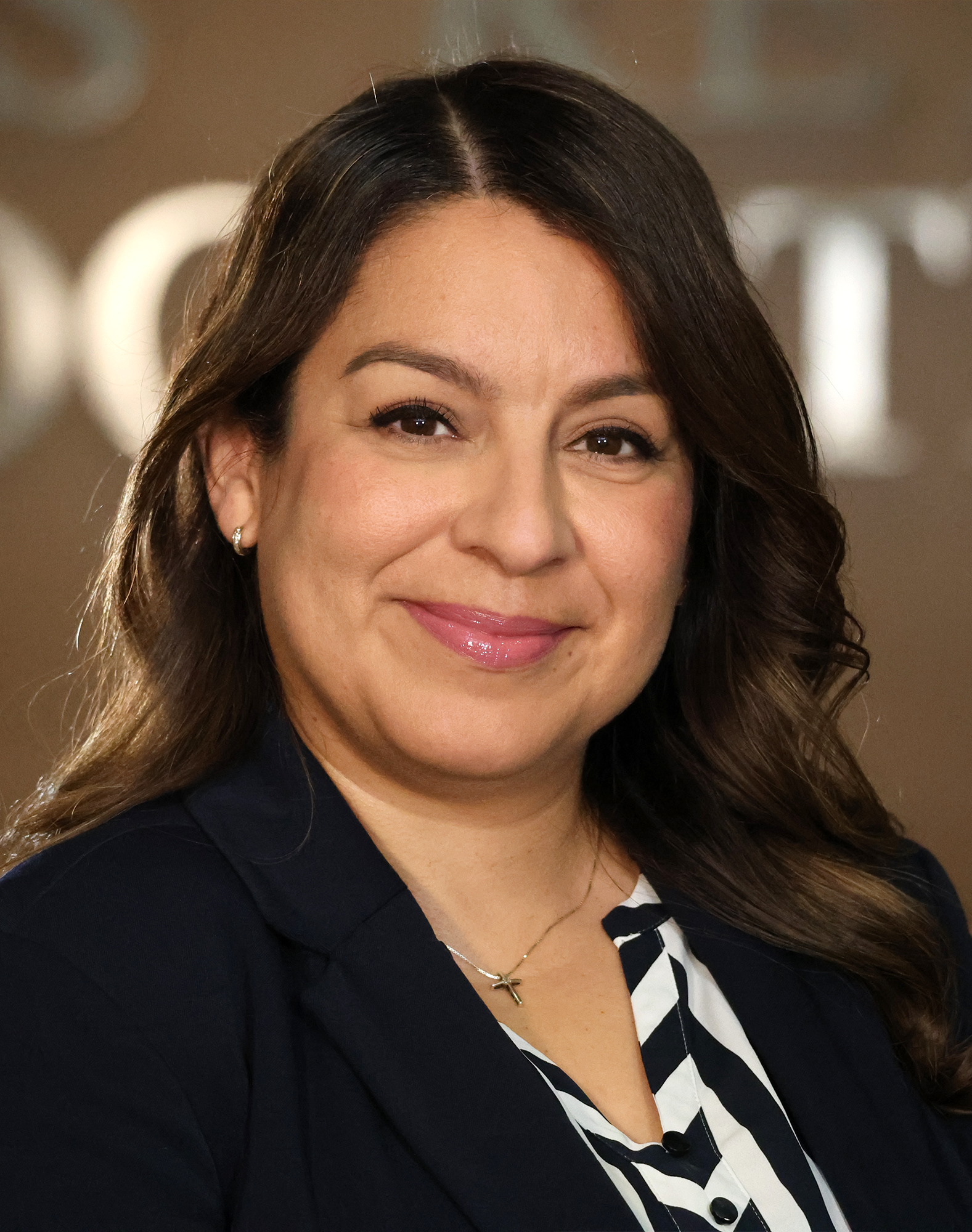 The image features Trustee Rocio Mosqueda smiling, dressed in a dark blazer over a striped shirt, with soft, styled hair.