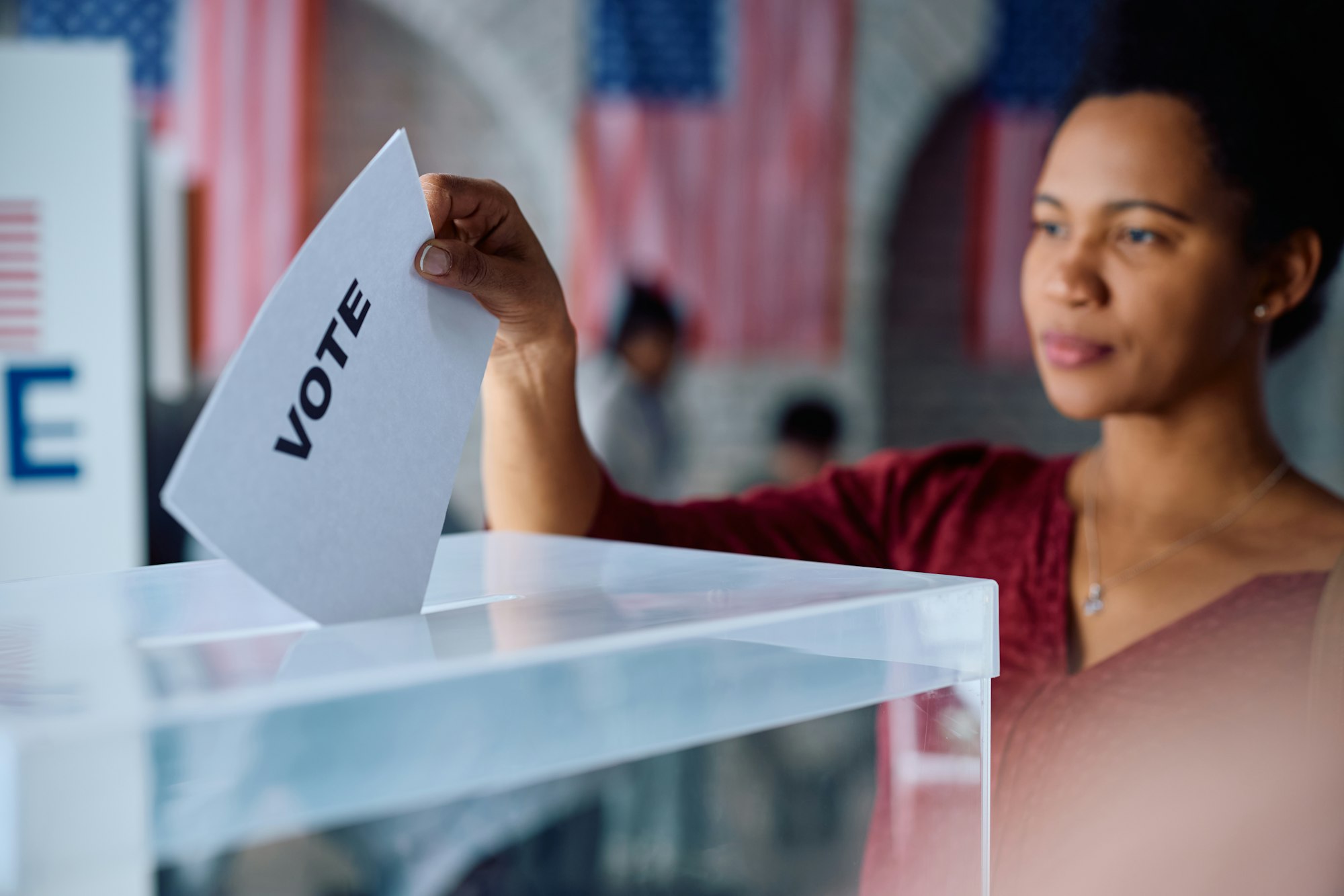 Adult woman dropping a voting ballot into a ballot box with American flags behind her