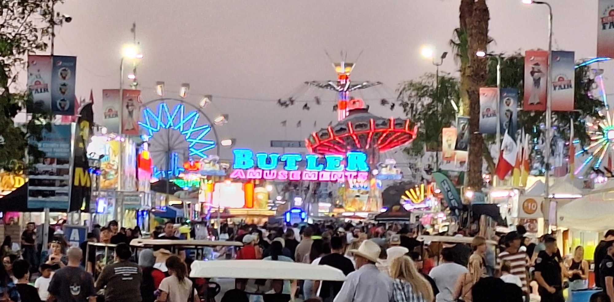 A bustling fairground with illuminated Ferris wheels, rides, "Butler Amusements" sign, banners, and a crowd of visitors enjoying the evening.