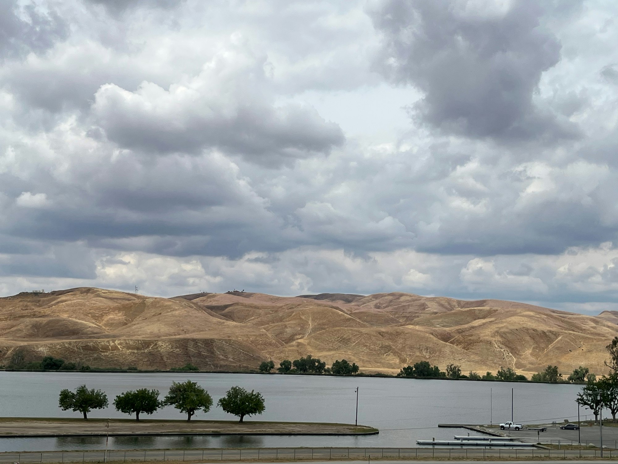 A lake, a dock, trees in a row, rolling hills, and a cloudy sky.
