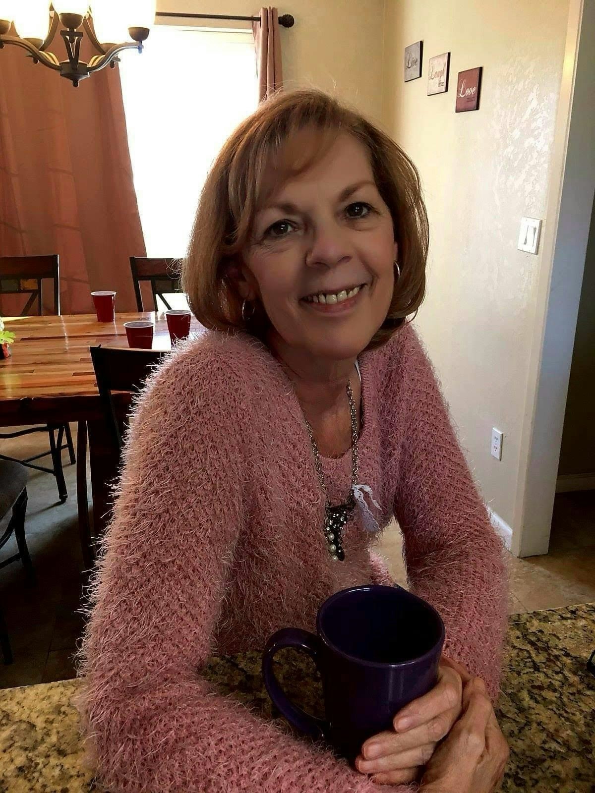 A smiling woman named Jan sitting at a table with a mug, a big warm smile, wearing a pink sweater, in a cozy room.
