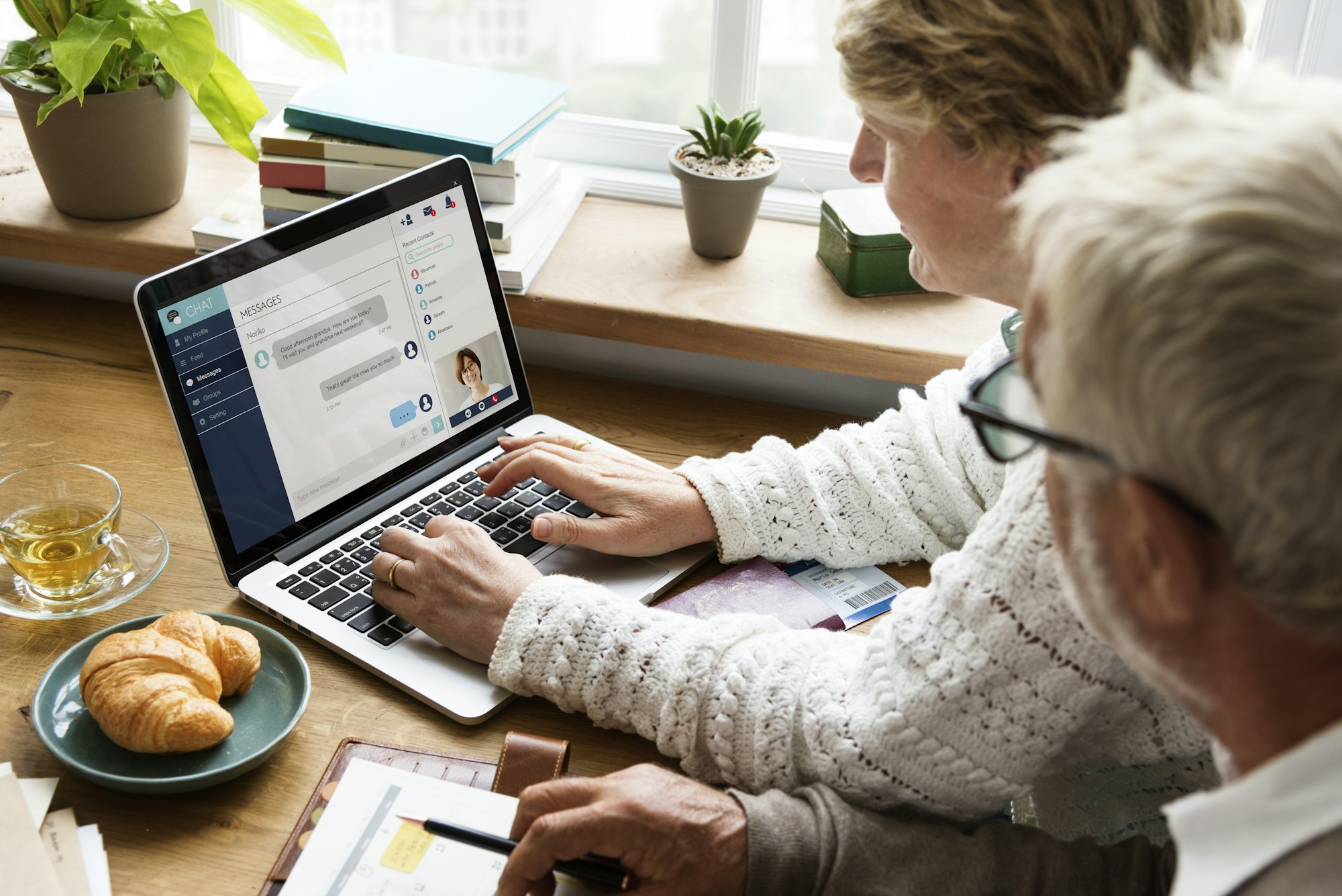 A person is chatting on a laptop with a beverage and croissant nearby, while another person observes. Books and a plant in the background.