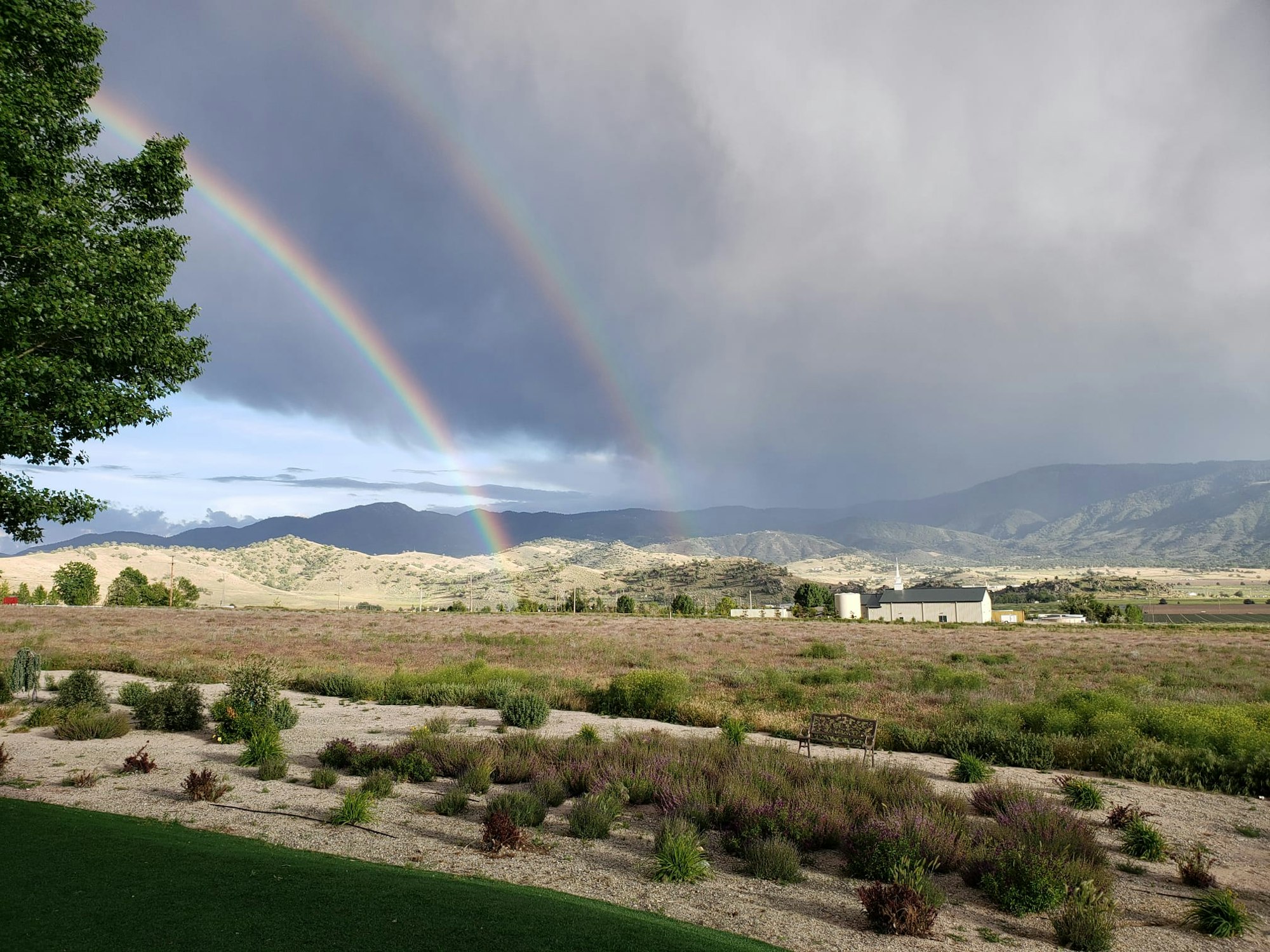 A double rainbow over a mountainous landscape with dark clouds, a garden in the foreground, and buildings in the distance.