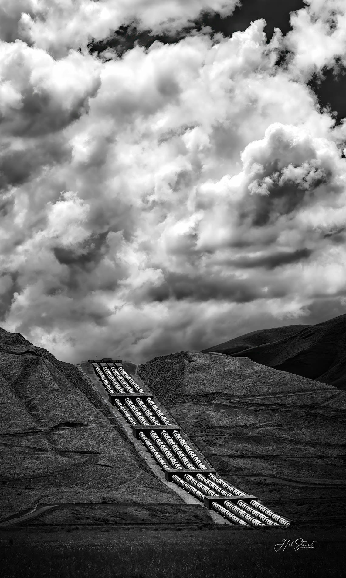 An industrial pipeline runs down a barren hillside under a cloudy sky, captured in black and white.