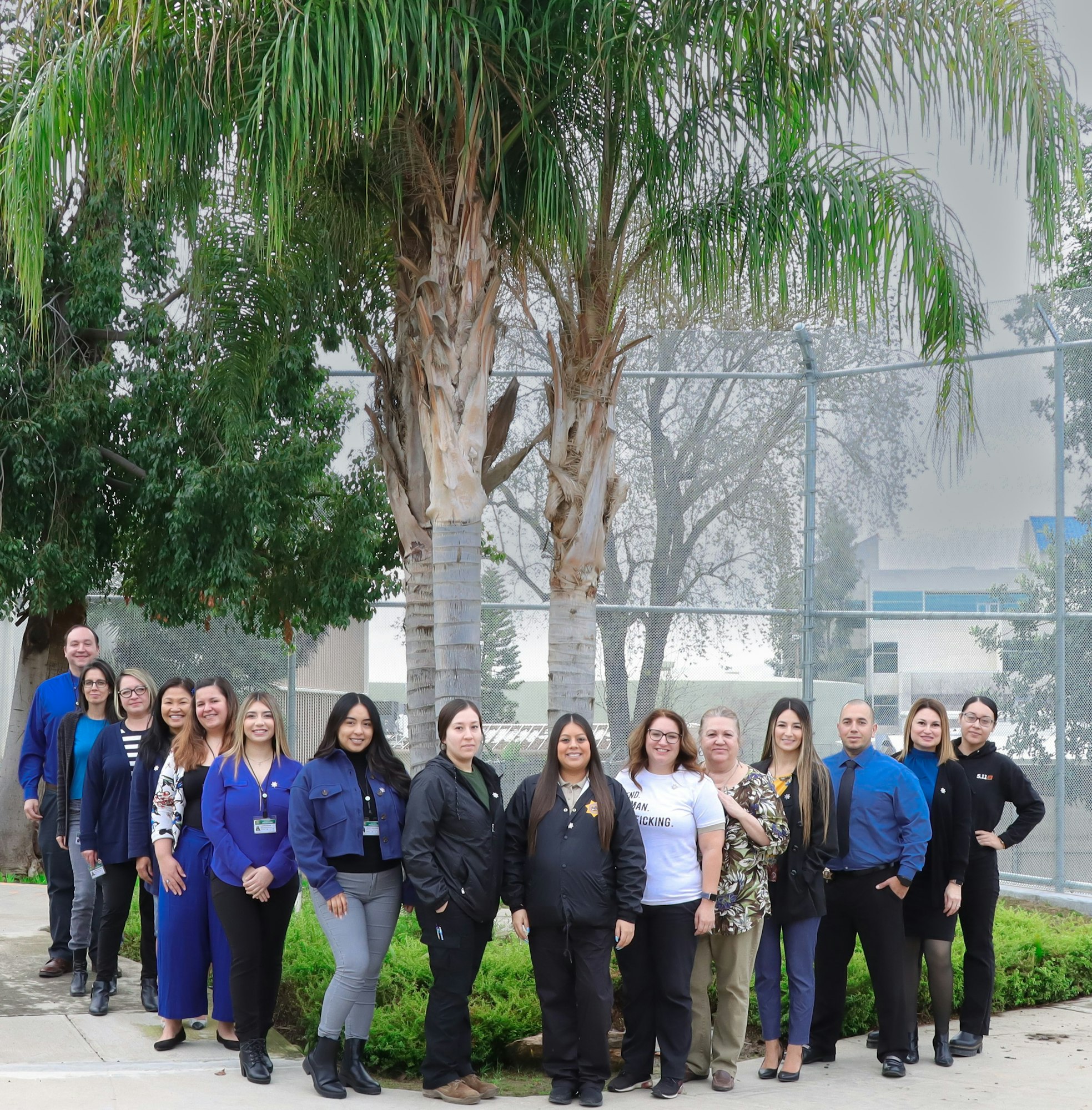 A group of people posing for a photo outdoors, with trees and a fence in the background.