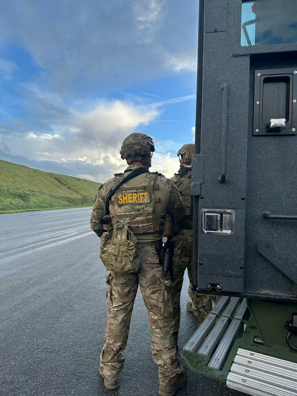 Soldiers in camouflage with "SHERIFF" patches near an armored vehicle under a cloud-filled sky.