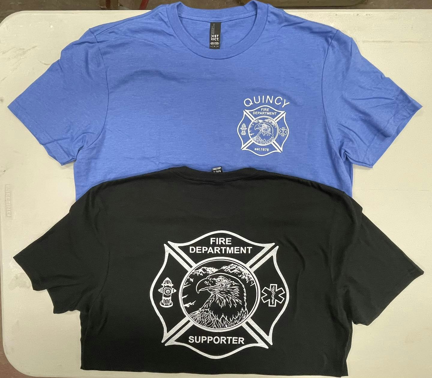 Two T-shirts with fire department logos, one blue on top and one black beneath, displayed on a flat surface.