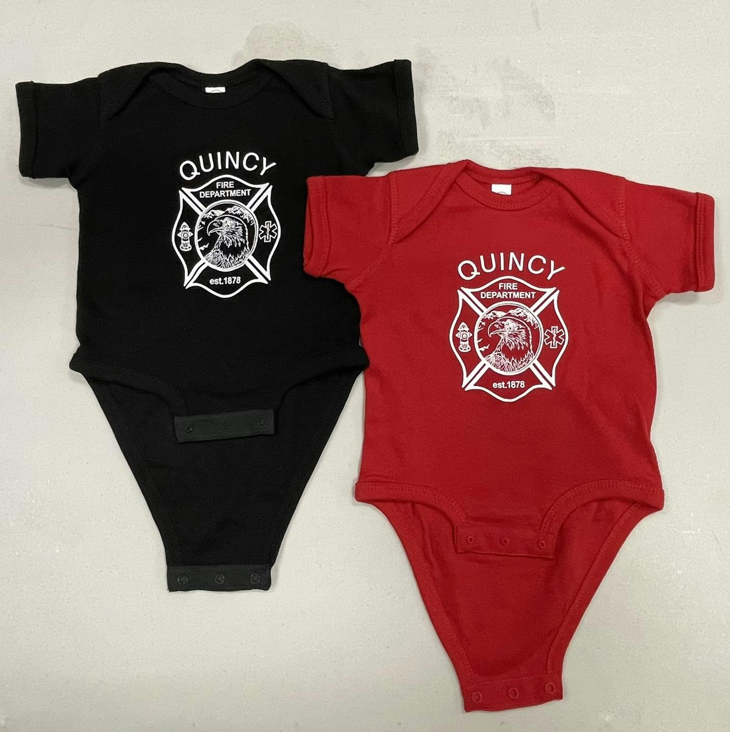 Two baby bodysuits, one black and one red, with "Quincy Fire Department" logos.