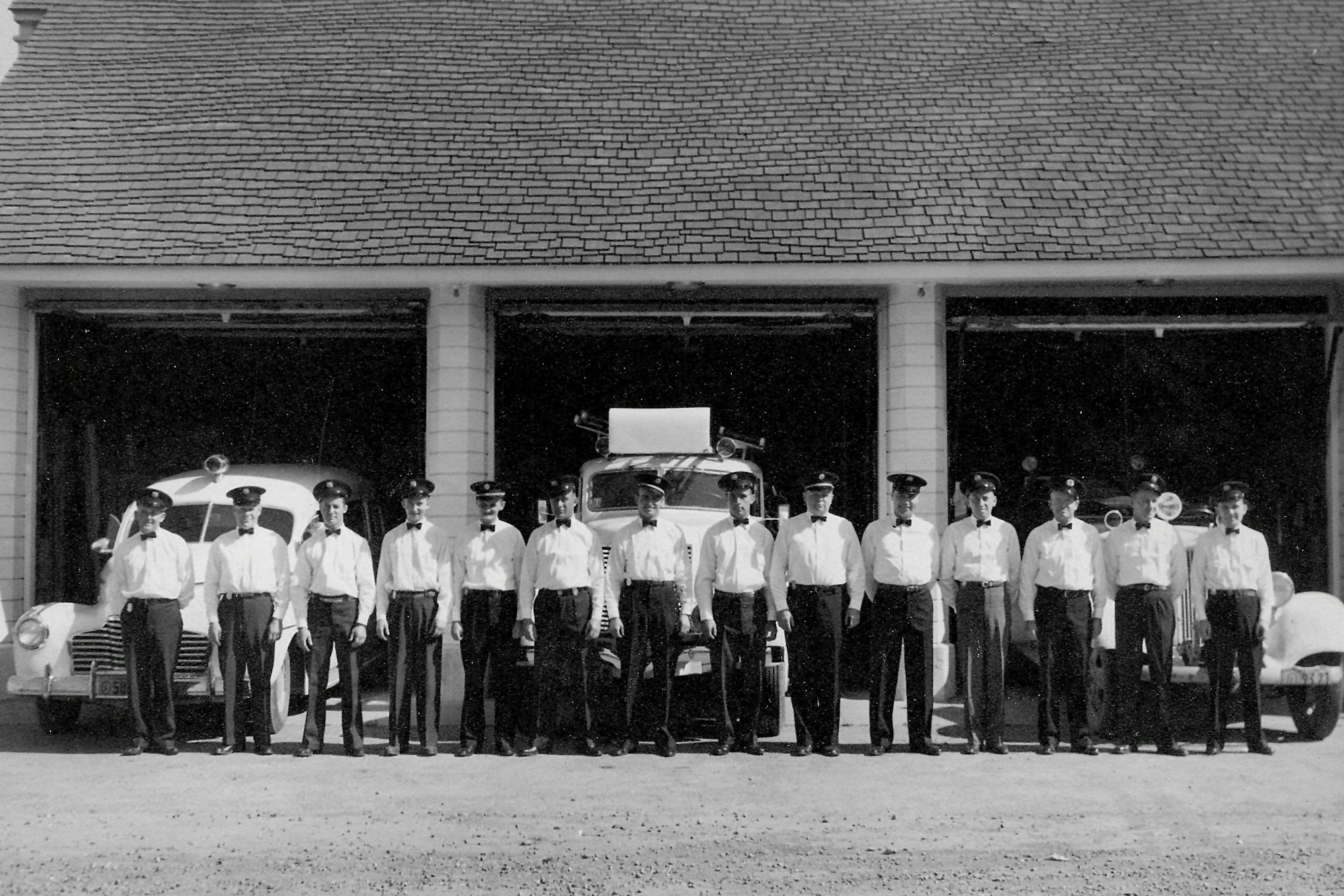 A group of uniformed firefighters standing in front of a fire station with fire trucks inside the garages.