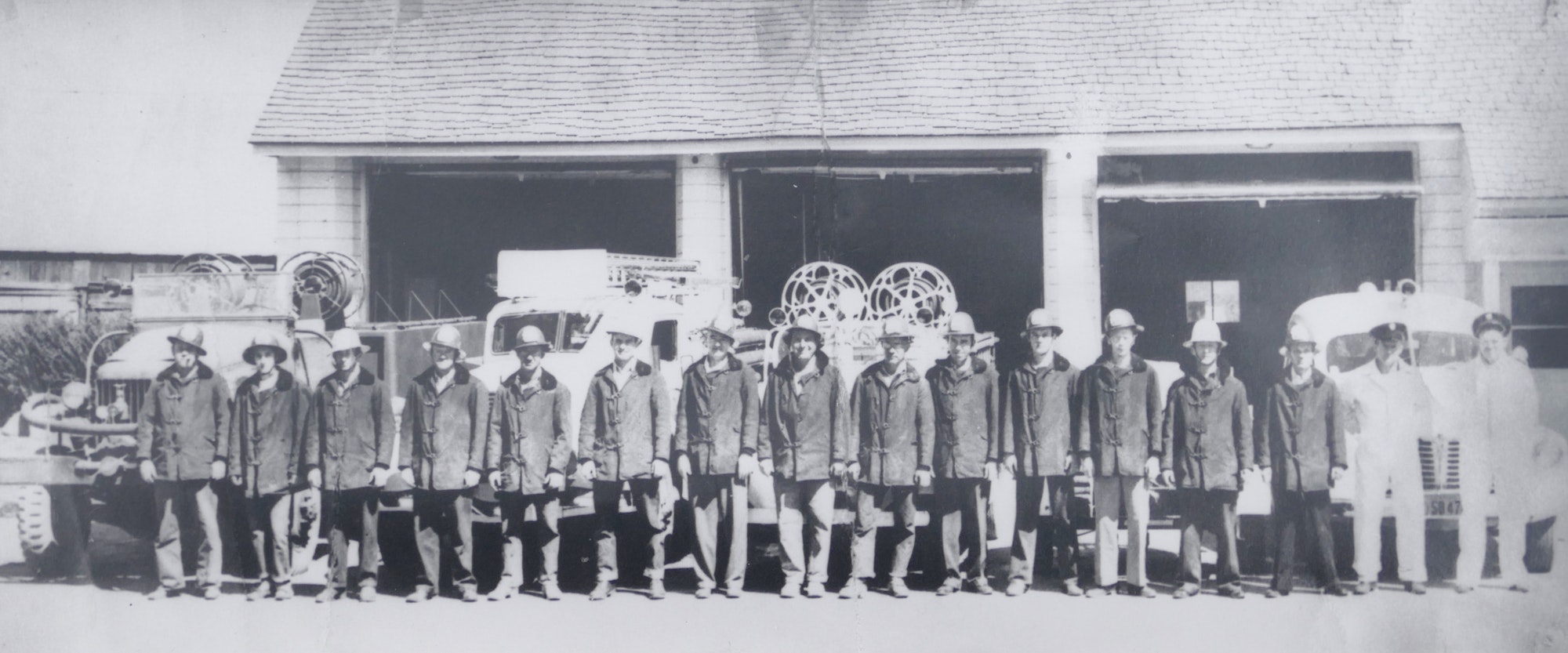 A group of firefighters in uniform standing in front of a fire station 1 in 1947 with fire trucks in the background.