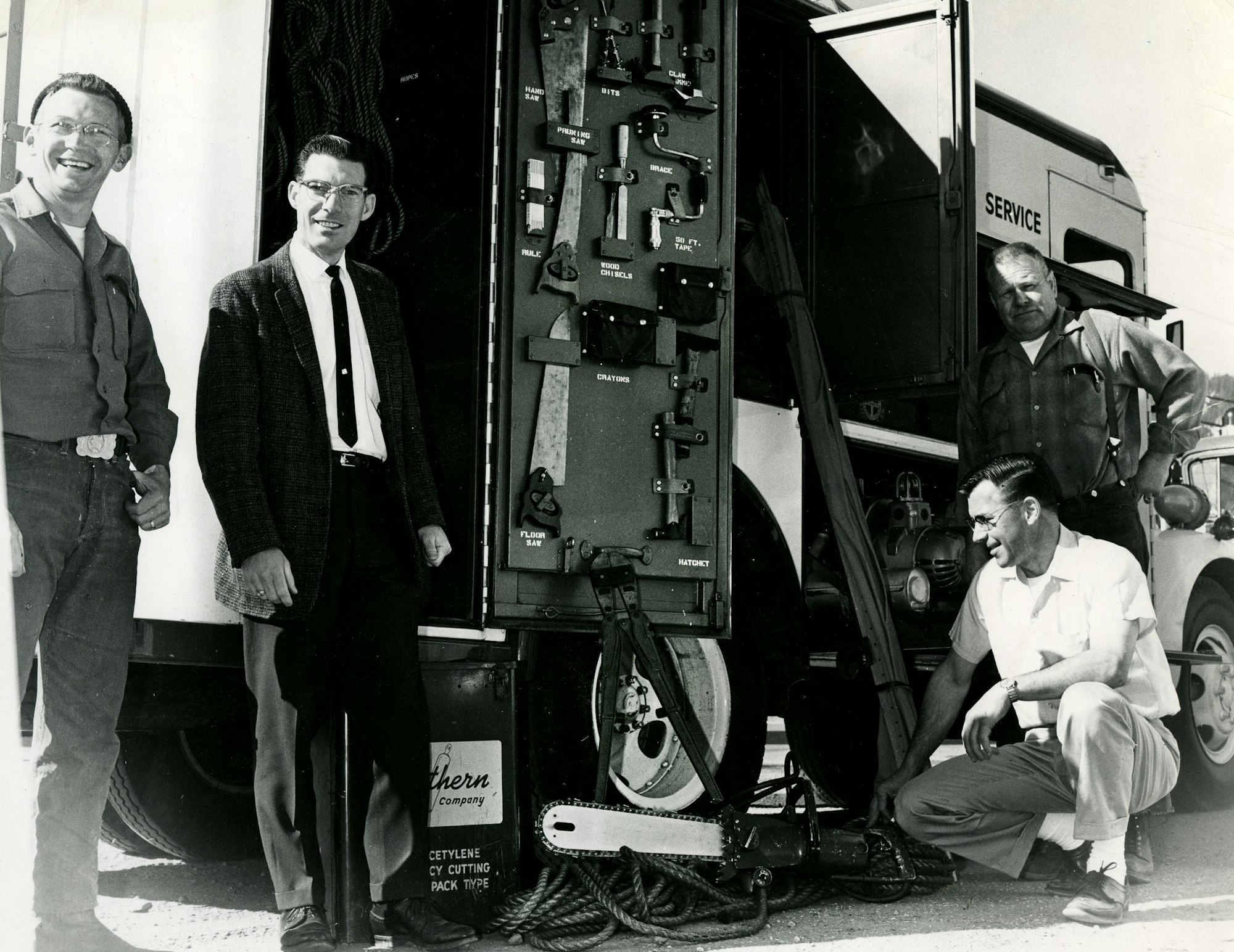Four men stand and kneel by a Civil Defense Rescue Rig service truck with tools on display.