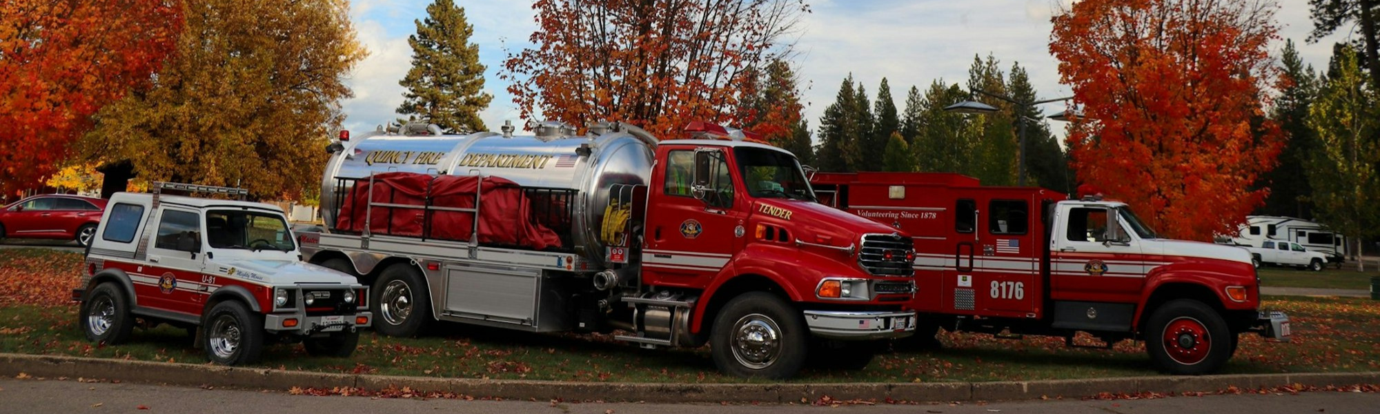 Three red fire department vehicles are parked in front of colorful autumn trees.