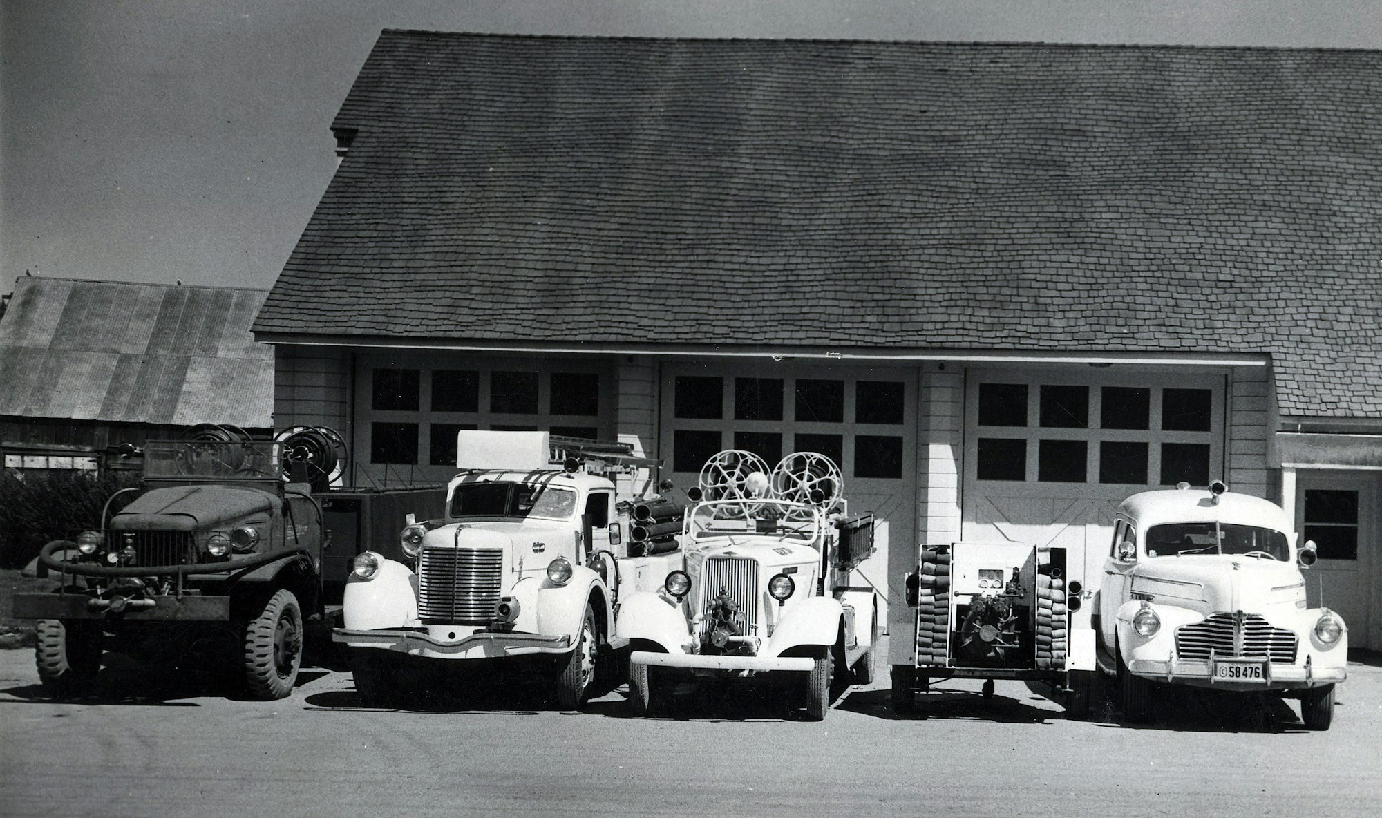 Five vintage fire trucks are parked in front of a fire station with a shingled roof.