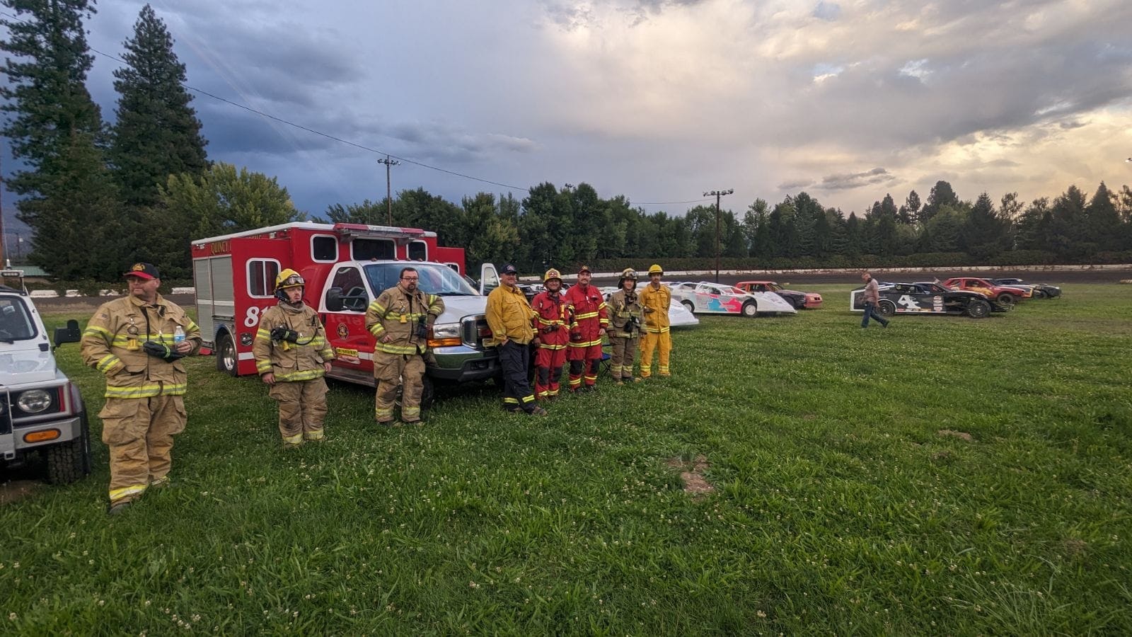 Rescue engine 8151 with Firefighters in gear stand near emergency vehicles and race cars on grass, under a cloudy sky.