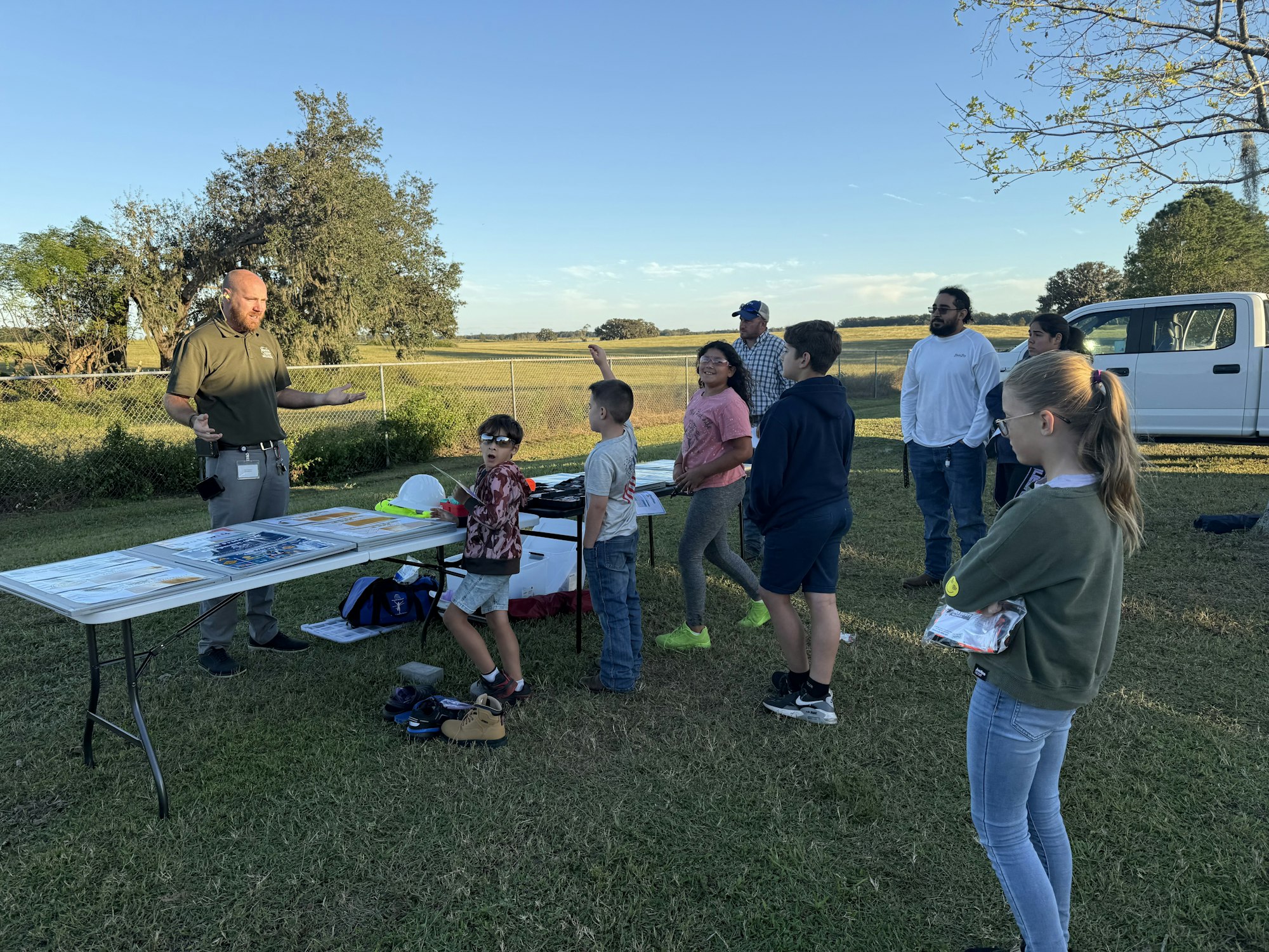 Group of people outdoors, possibly at an educational event, with a man presenting and various items on a table.