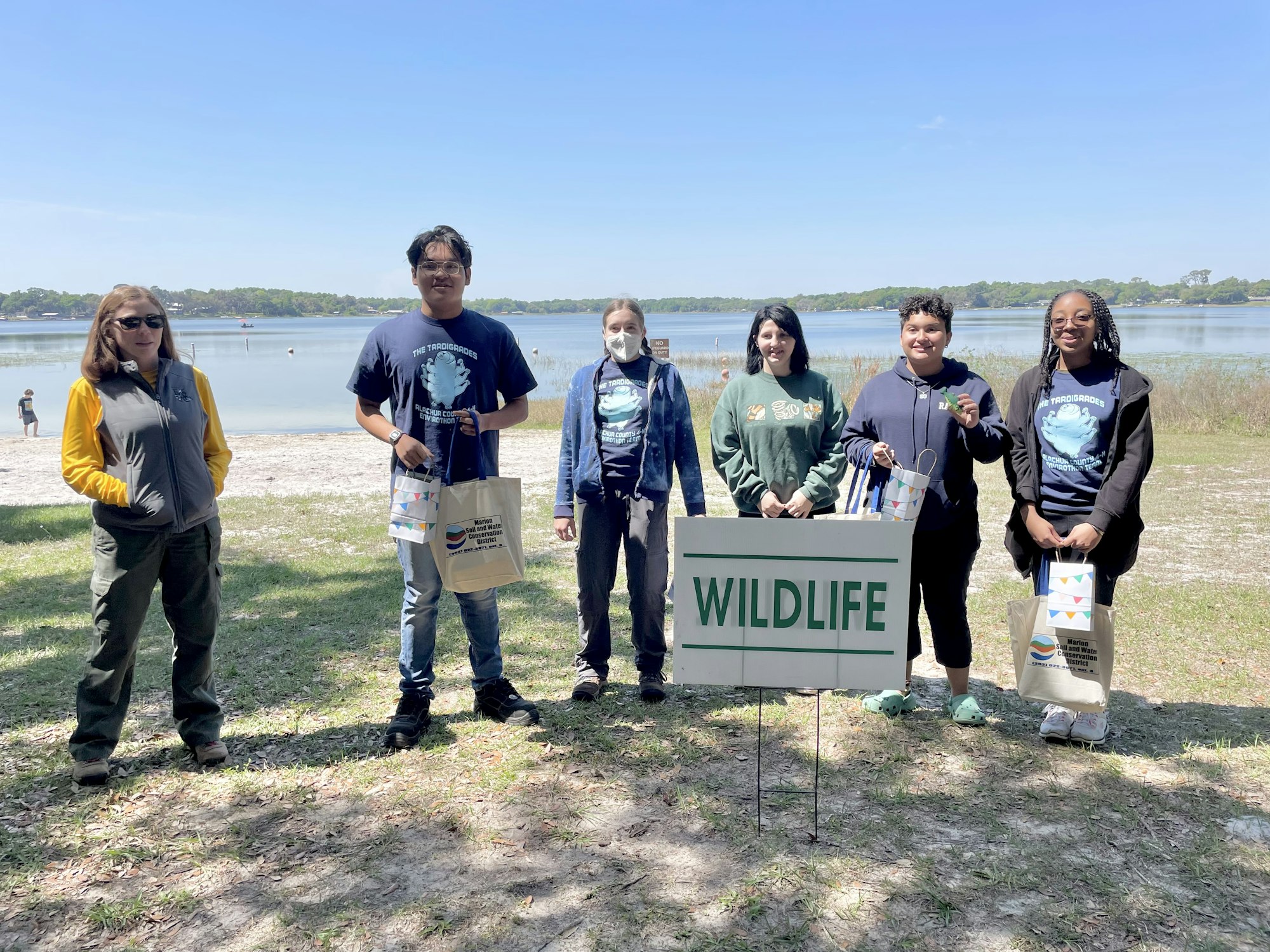 Group of people by a lake with a "WILDLIFE" sign, holding bags and wearing outdoor clothes.