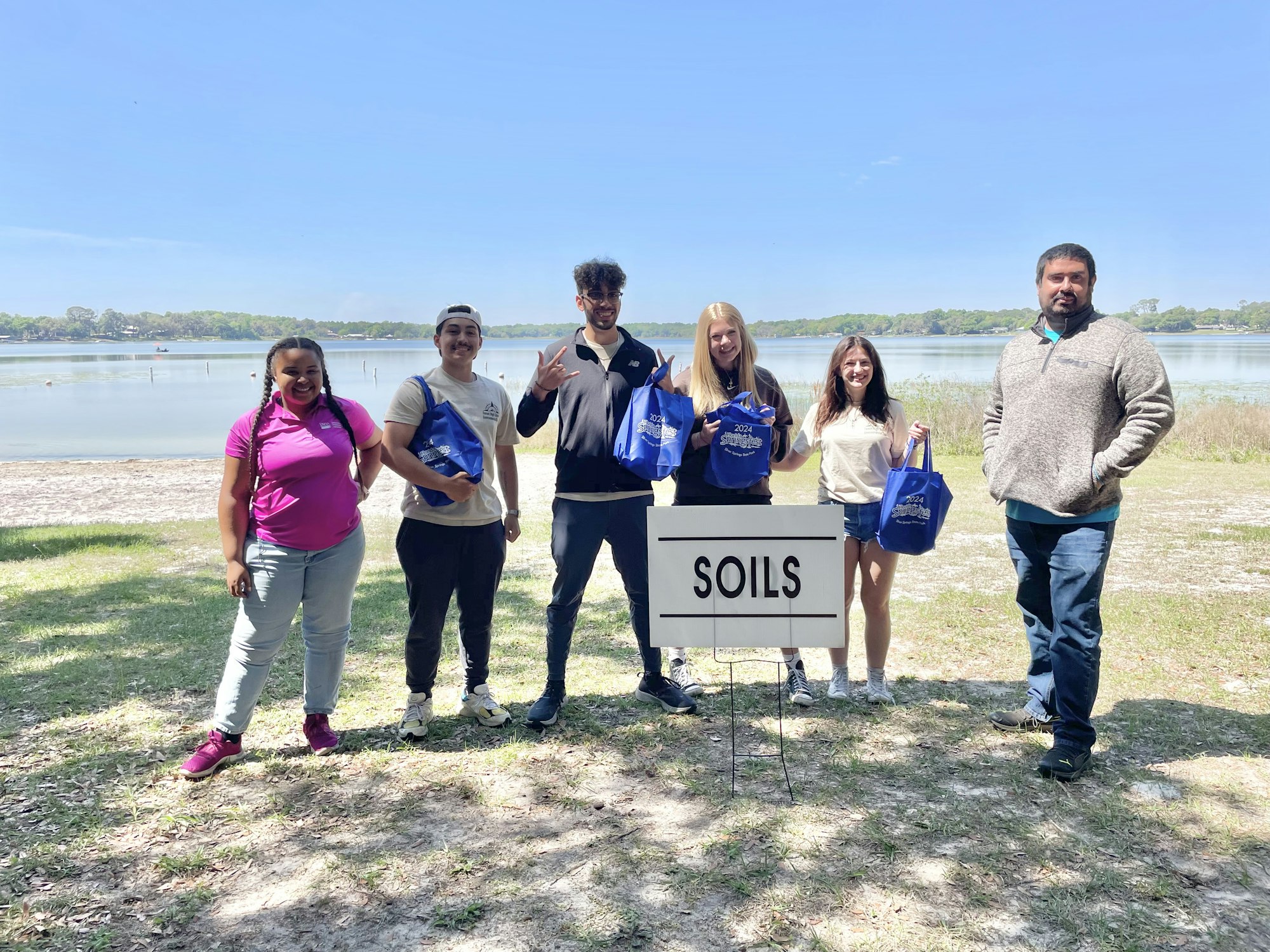 Six people posing by a "SOILS" sign near a lake, holding blue bags, likely at an outdoor event or class.