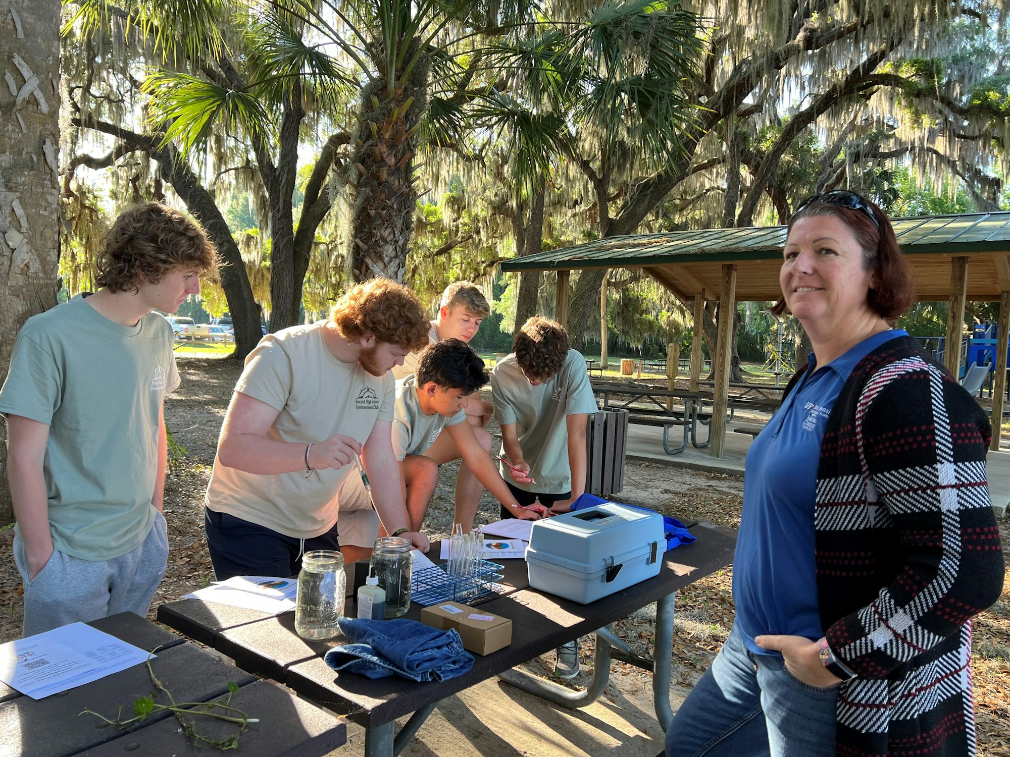 A group of people engaged in an outdoor activity with scientific equipment on a table, surrounded by trees.