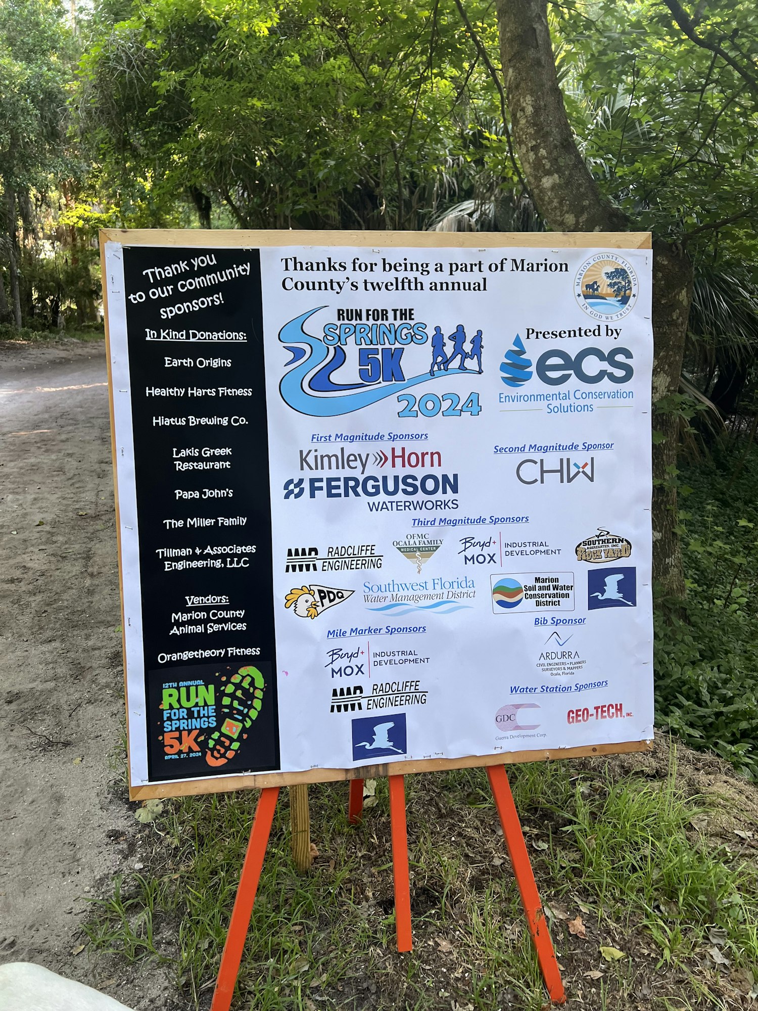 A sign with multiple sponsor logos for an annual Springs 5K run event, displayed vertically outdoors.