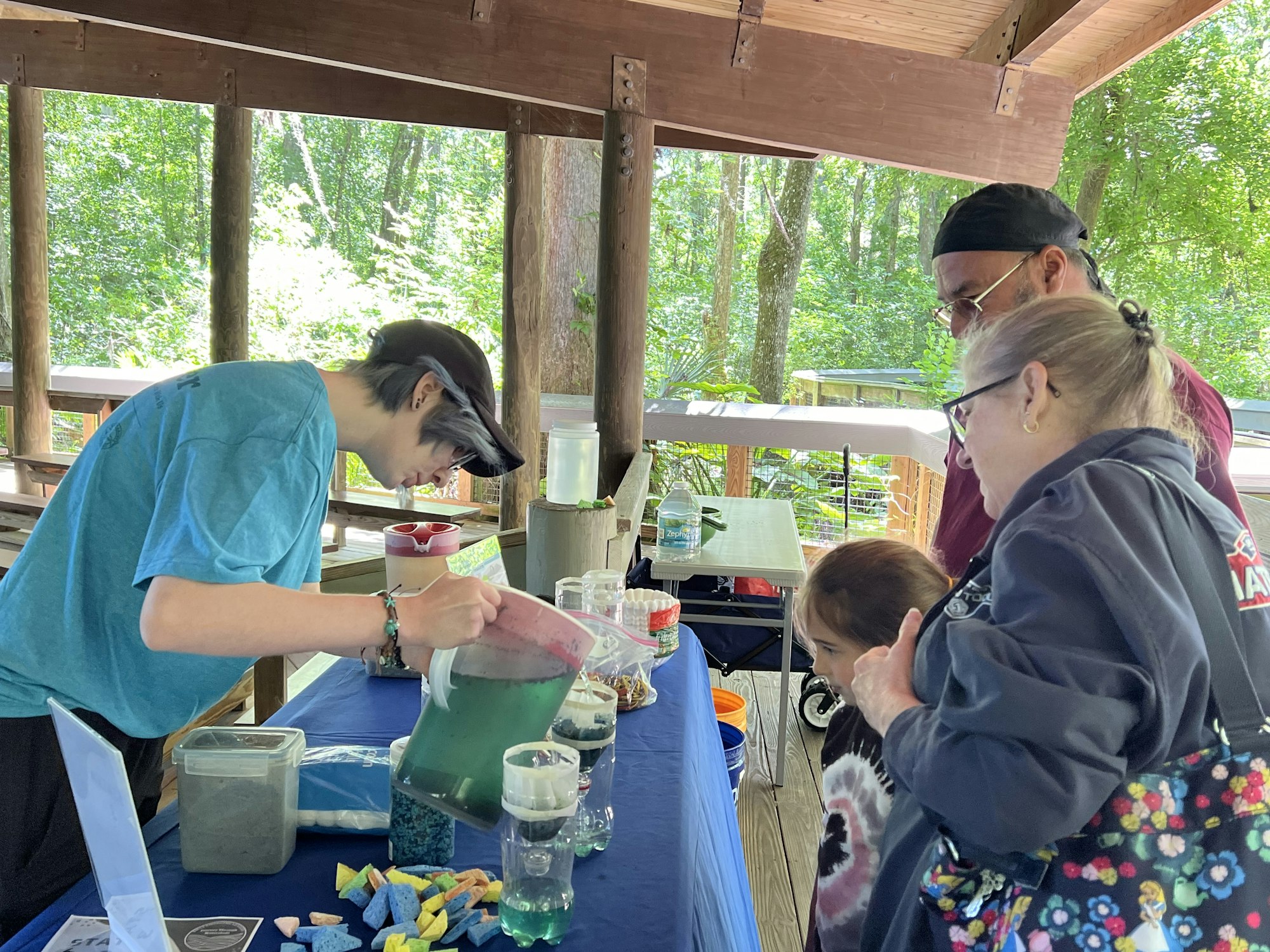 People at a table with various items, possibly at an educational or craft activity outdoors.