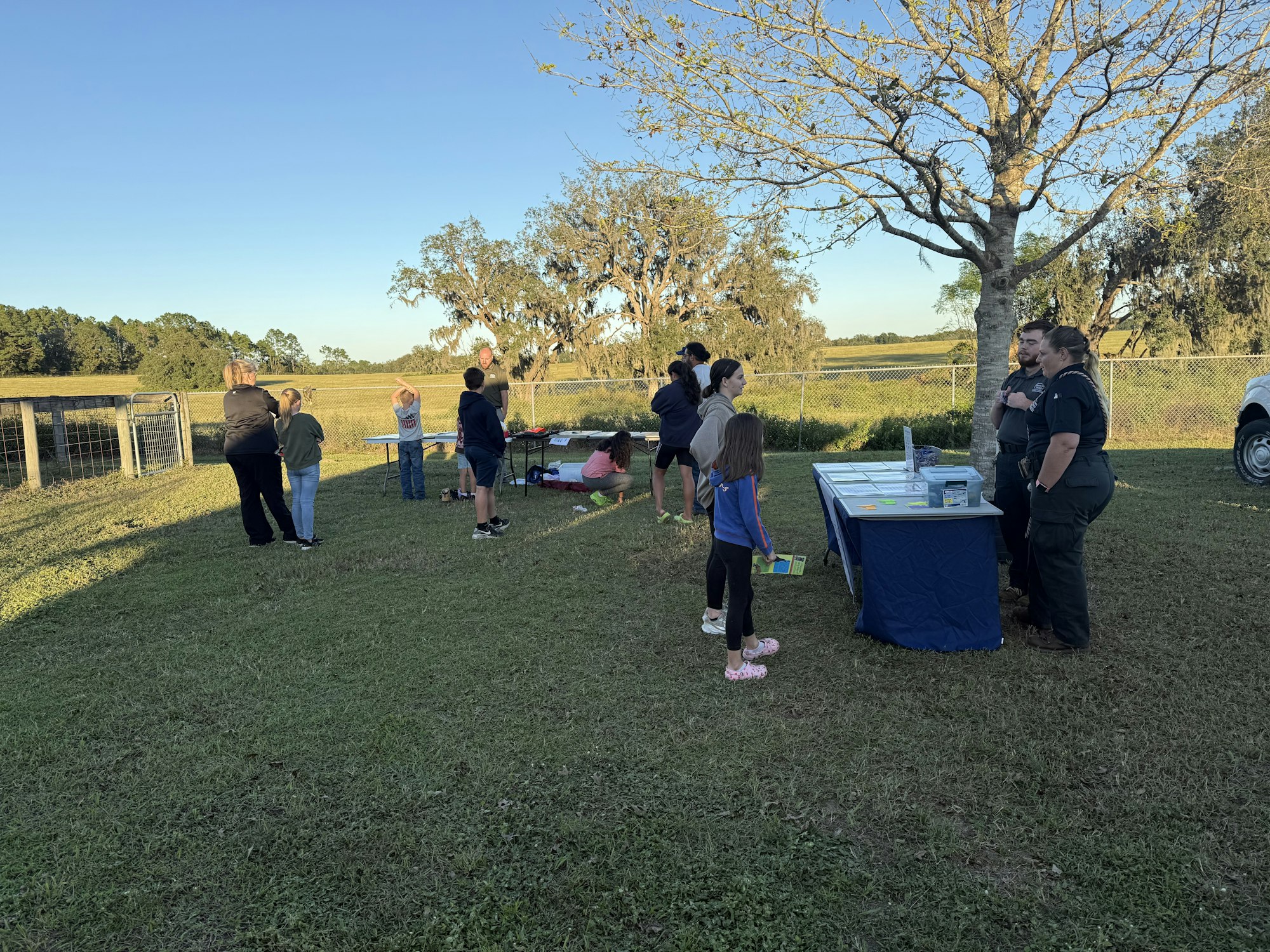An outdoor scene with people gathered around a table, some talking to uniformed officers, in a grassy area with trees and sunlight.