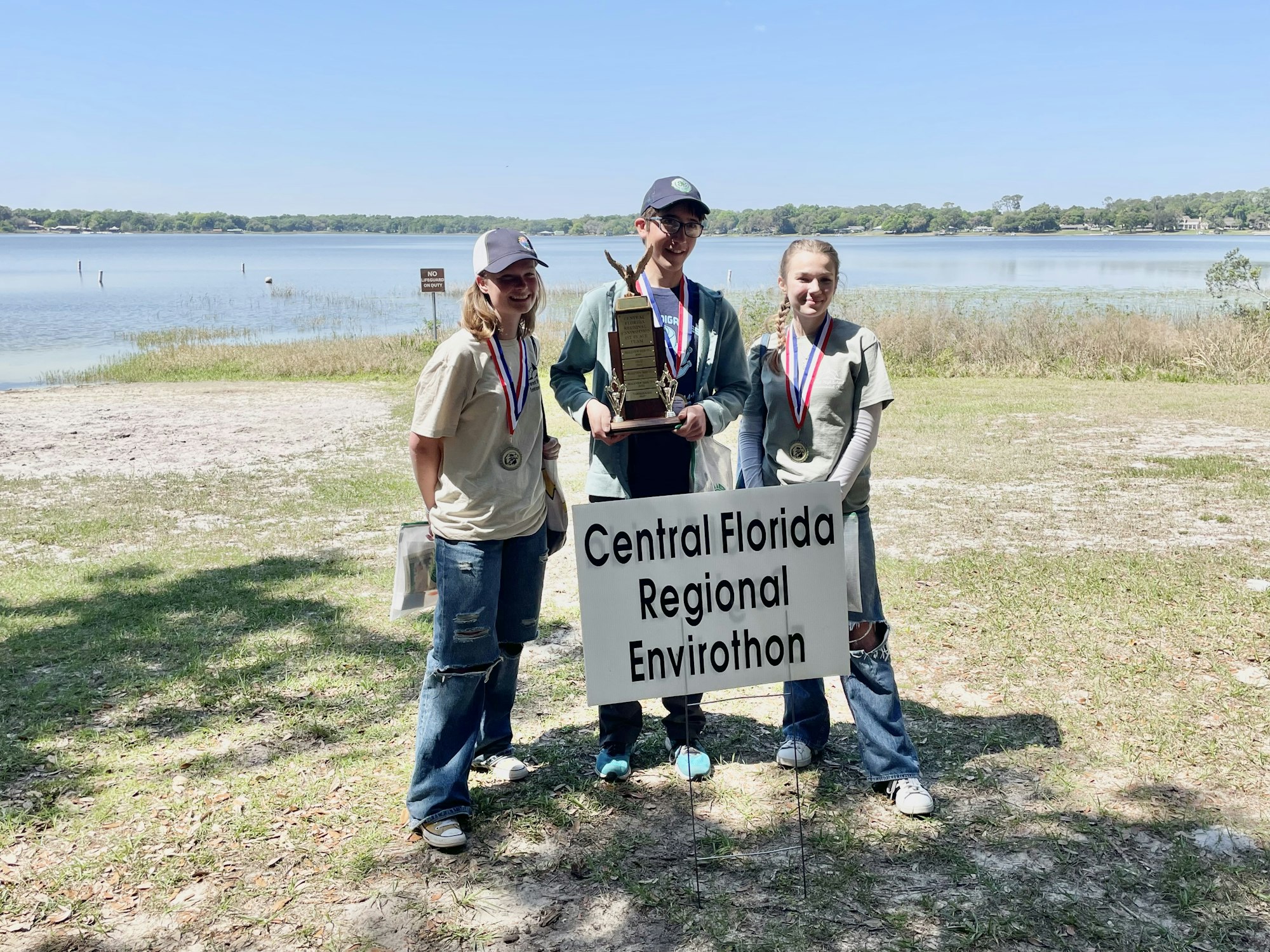 Three individuals with medals and a trophy, standing by a sign "Central Florida Regional Envirothon" near a lake.