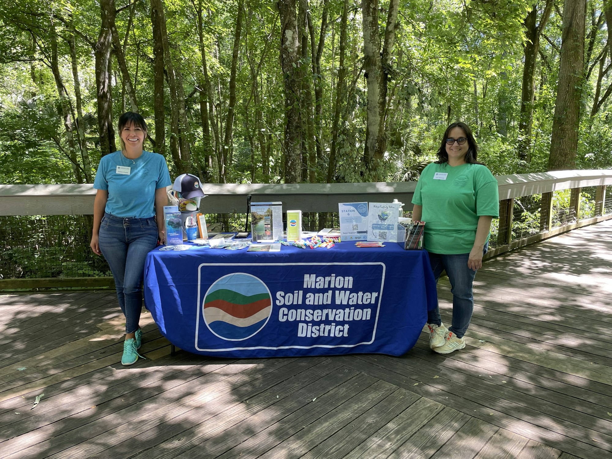 Two people stand by an informational table with banners in a wooded outdoor area.