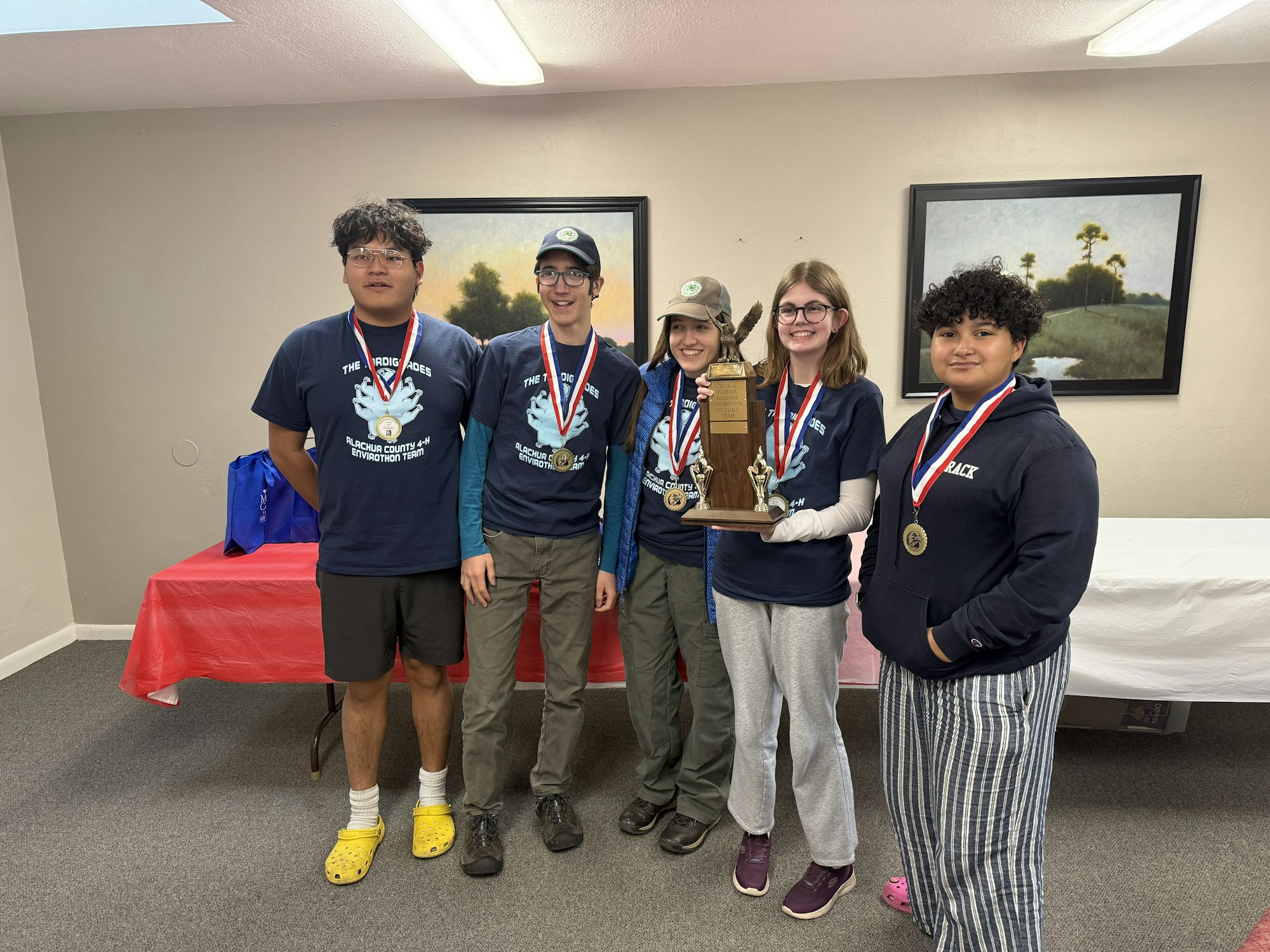 Five people wearing medals hold a trophy, standing indoors against a beige wall with framed artwork.