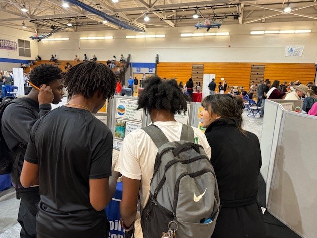 Students gather at a school event or fair with booths and attendees in the background.