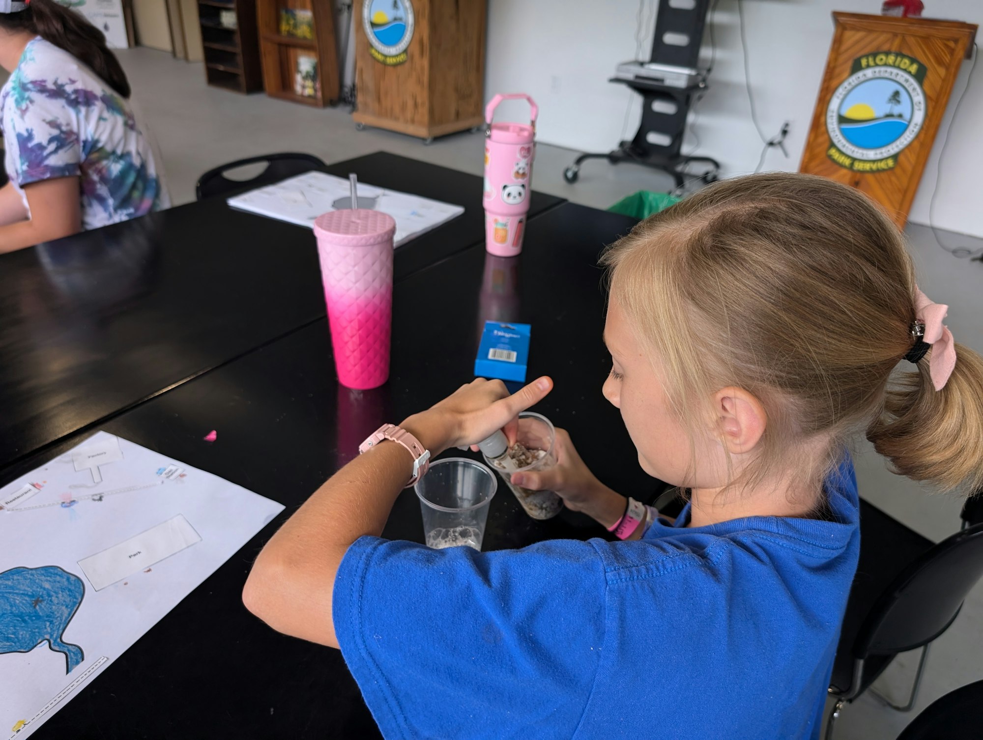 A child at a table with papers, drinking cups, in what appears to be a classroom setting.