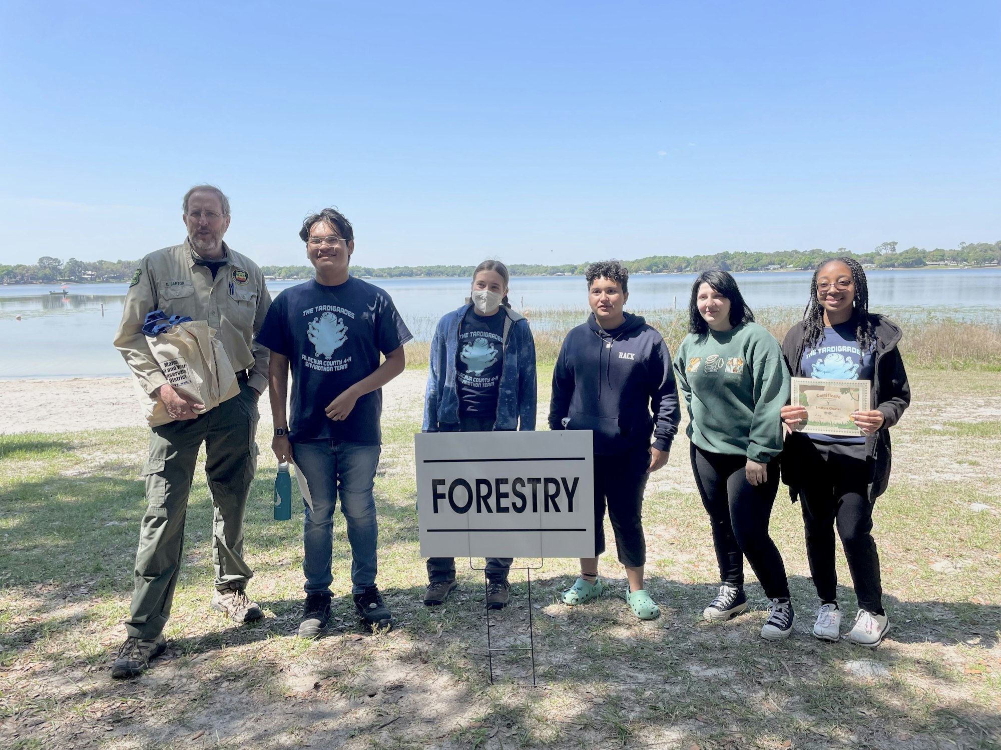 A group of six people posing by a "FORESTRY" sign, likely at an outdoor event or competition, with a lake in the background.