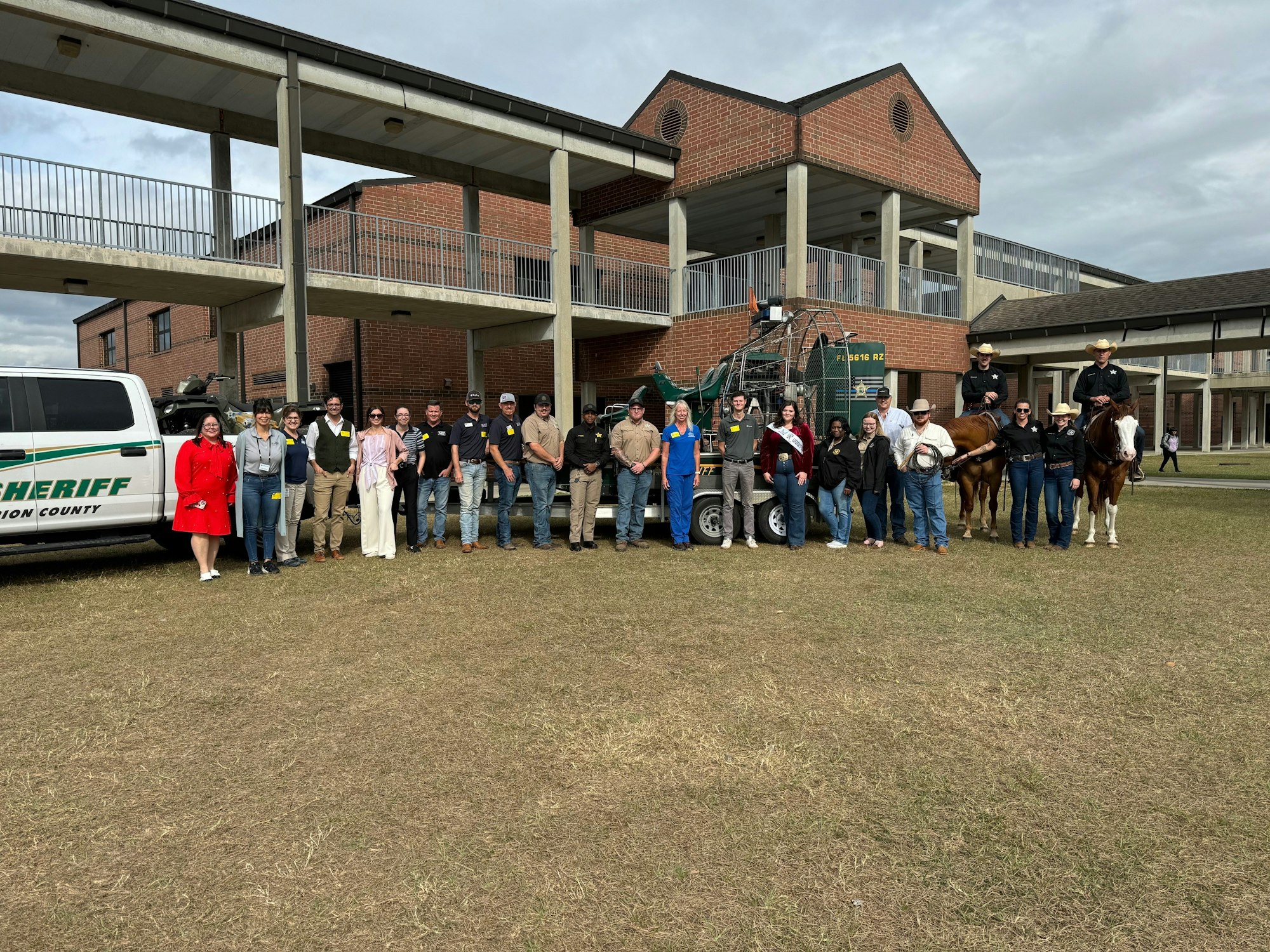 Group of people posing with horses in front of a sheriff's van and building.