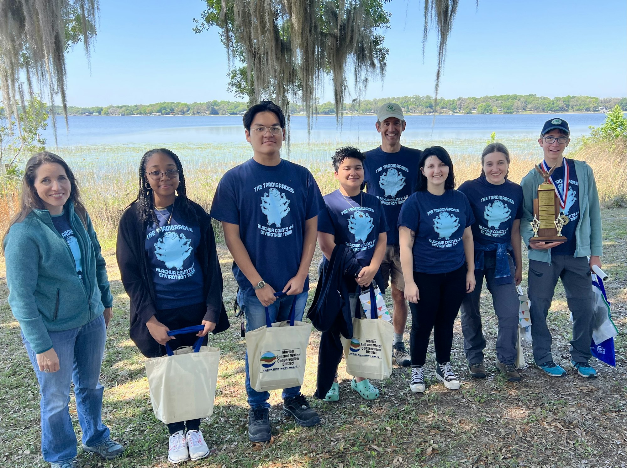 Group of people outdoors, some holding bags and a trophy, by a lake with Spanish moss-draped trees.