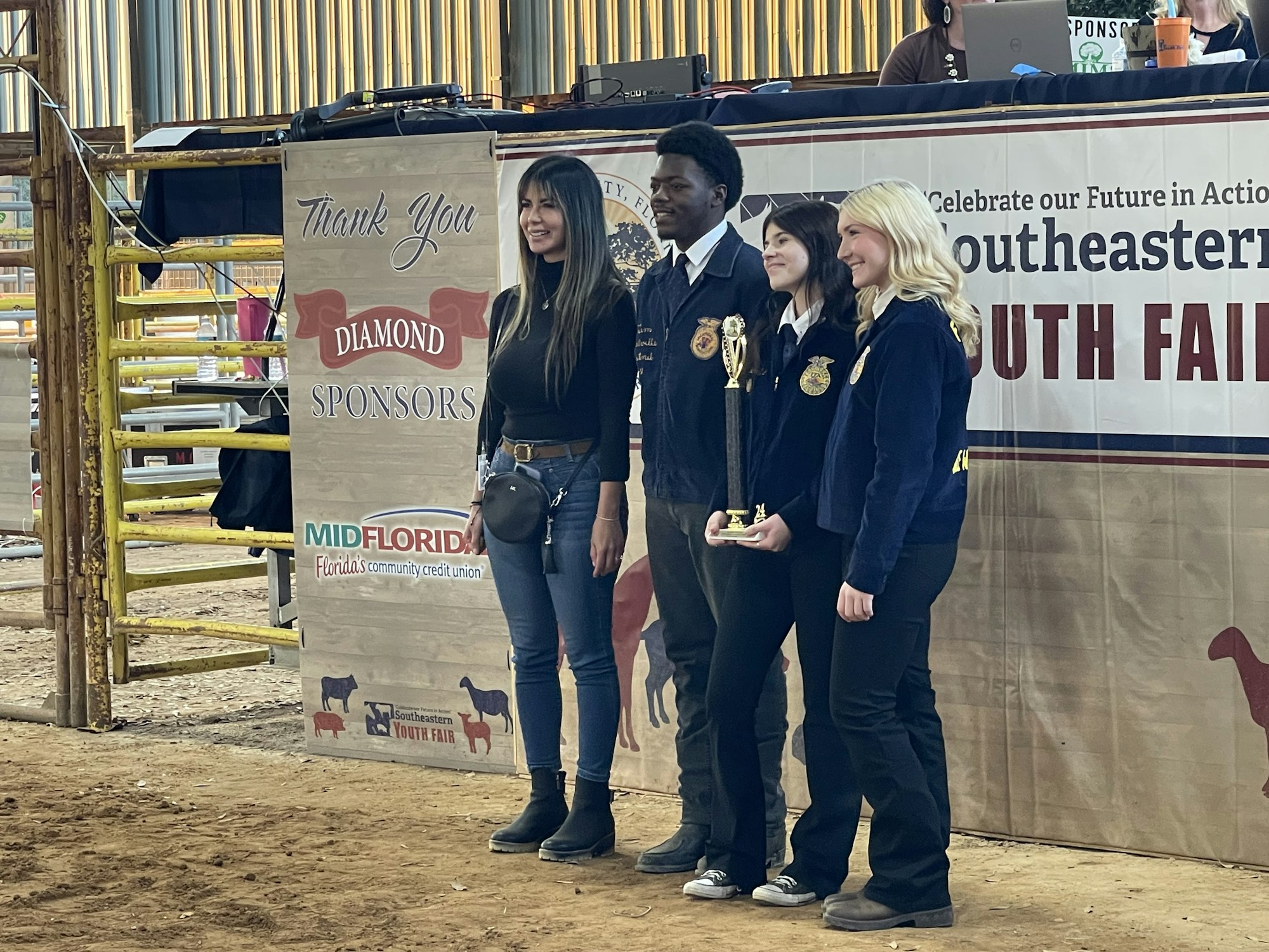 Three people at a youth fair event holding a trophy, with sponsor banners in the background.