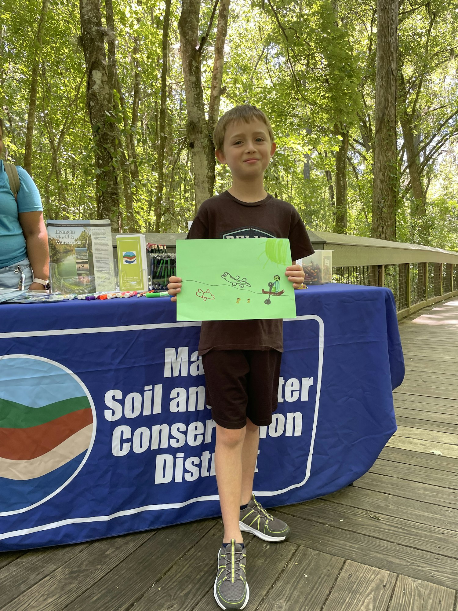 A child stands on a deck holding a drawing, with a "Soil and Water Conservation District" banner behind. Trees surround the scene.