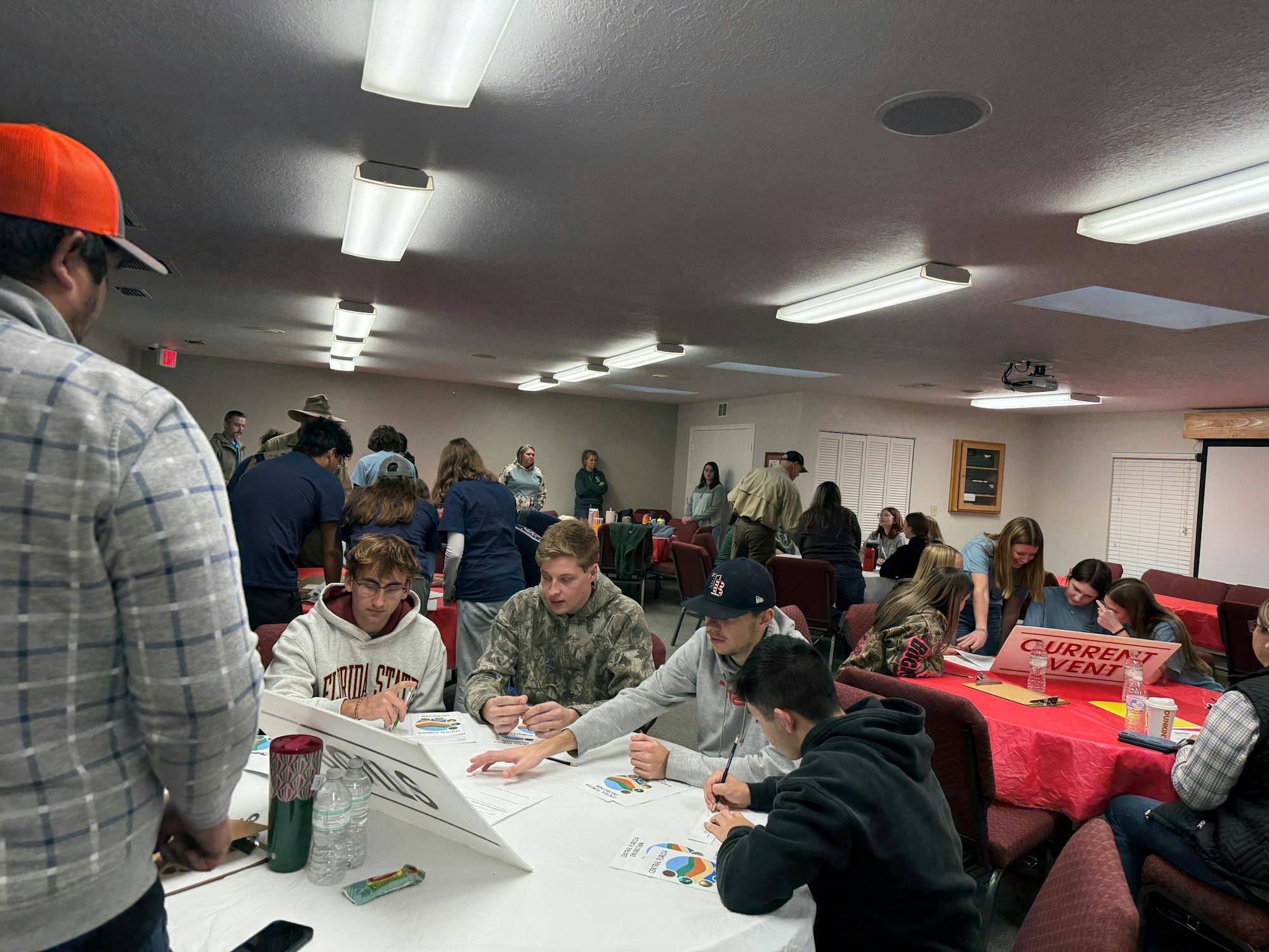 A group of people indoors, sitting at tables and engaging in activities or discussions—sign labeled "Current Events" visible.