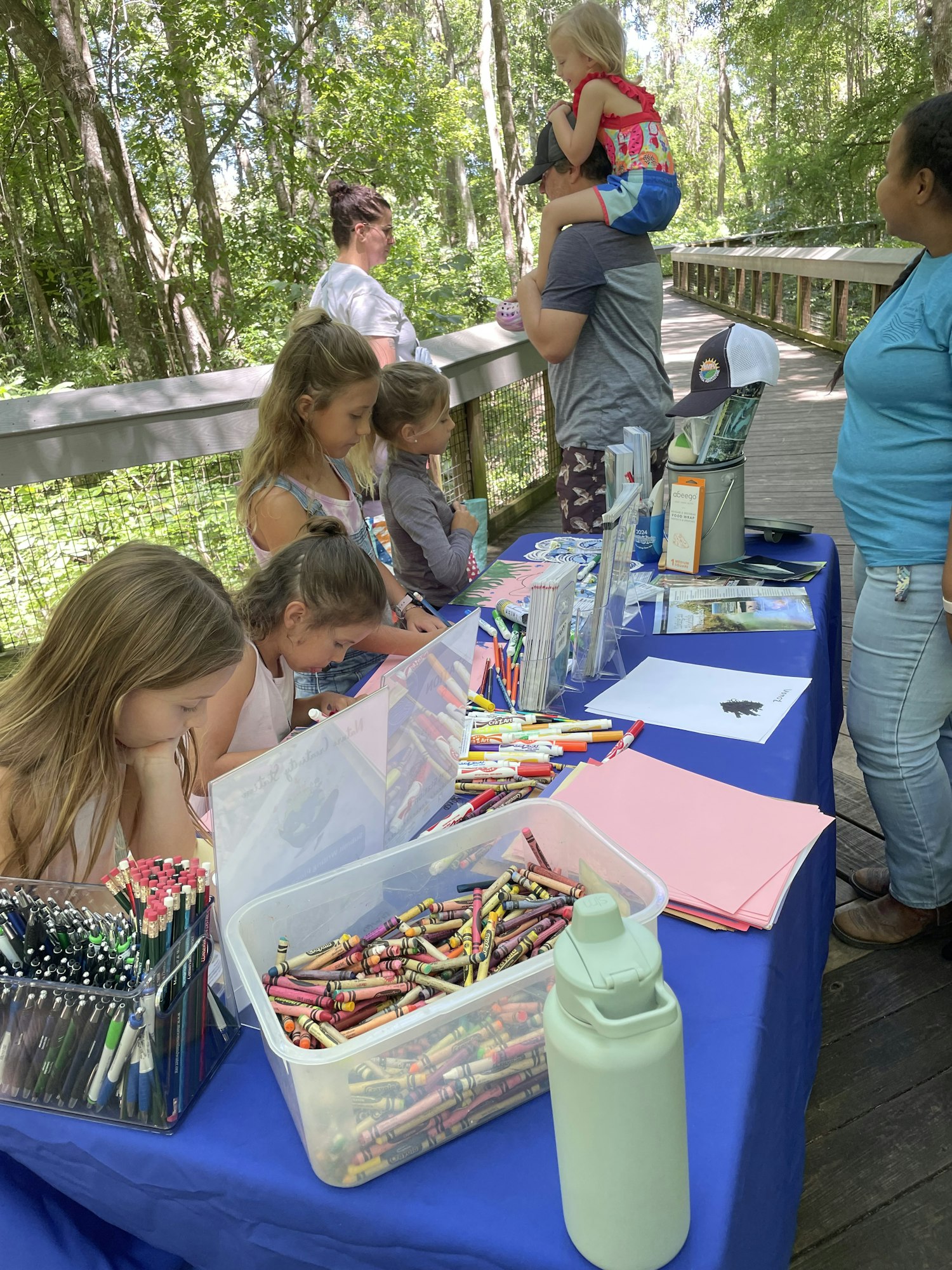 Kids at a craft table with papers and crayons in a wooded outdoor setting.