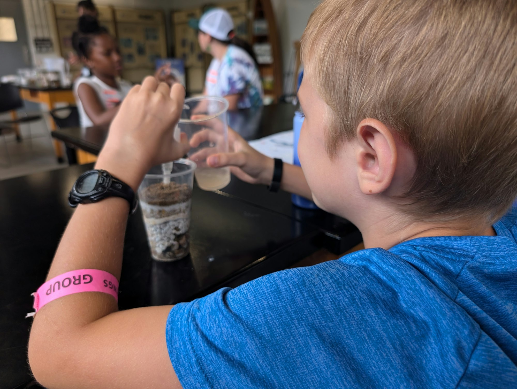 A child engaging in a hands-on activity, holding a clear cup with layers of soil, with others around in a casual setting.