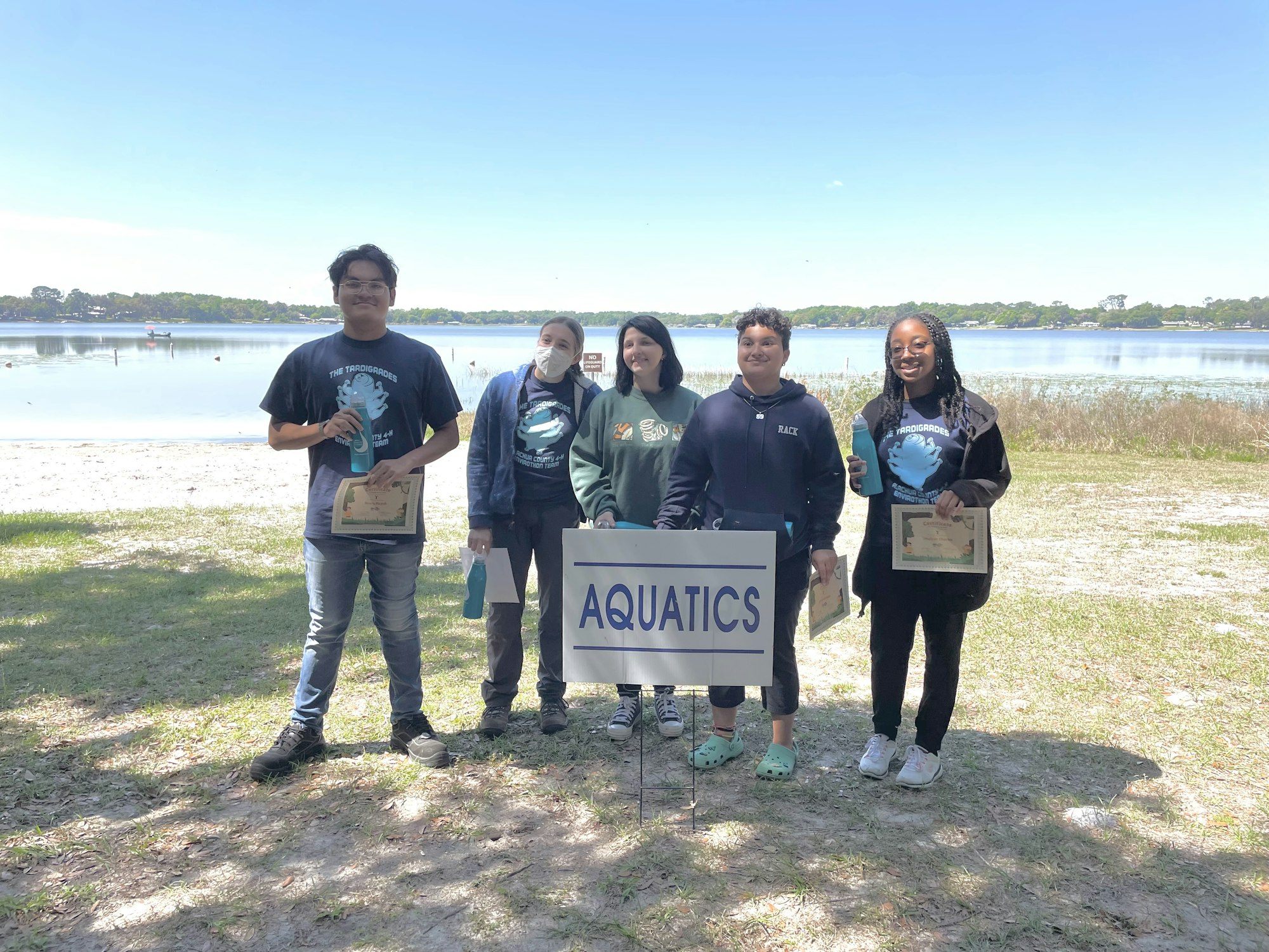 Five people by a lake with certificates, standing behind an "AQUATICS" sign.