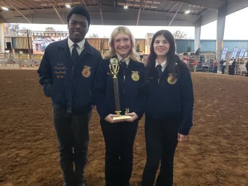 Three people in FFA jackets holding a trophy, standing in a livestock arena.