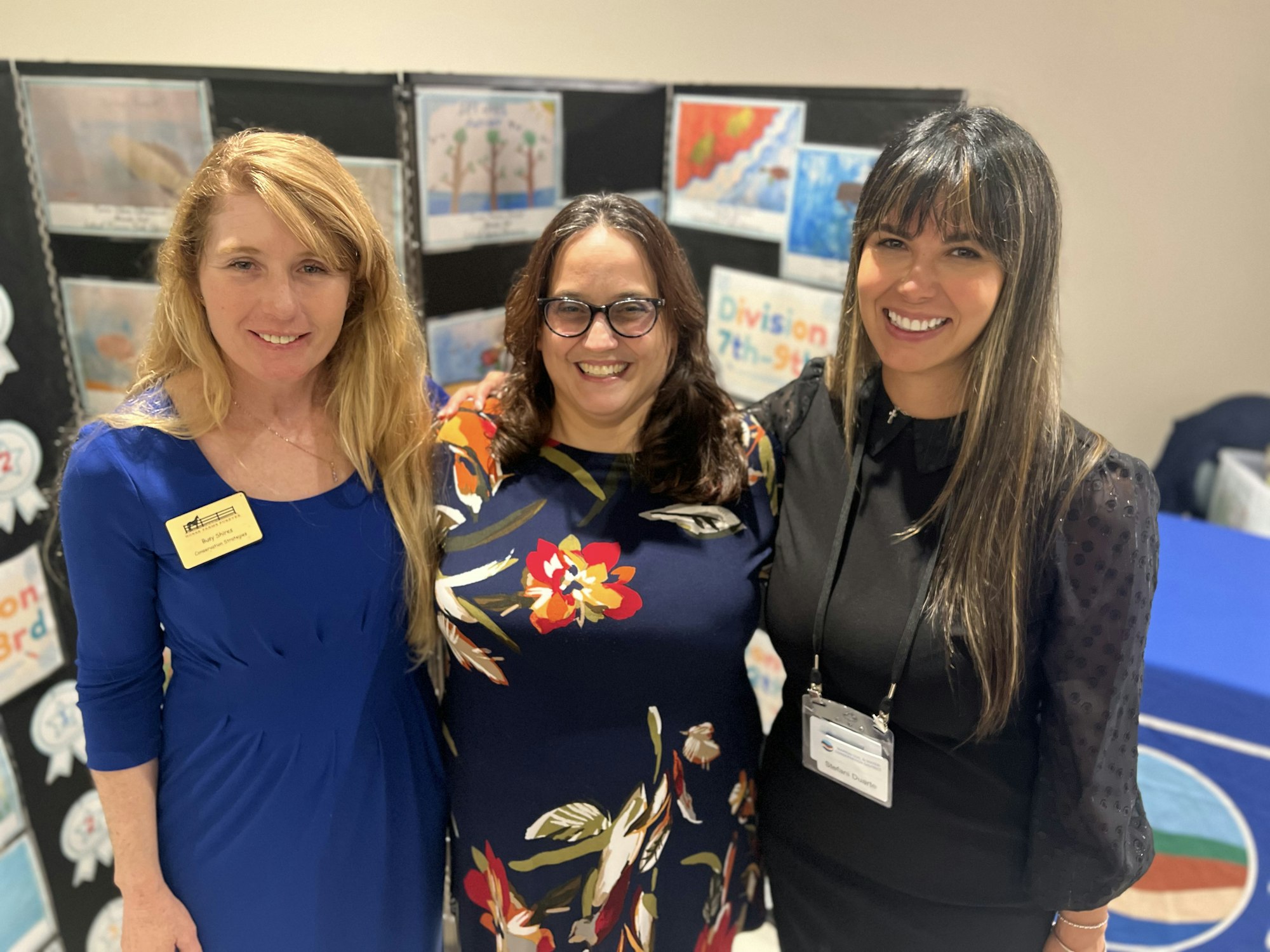 Three smiling women posing in front of a display board with artwork.