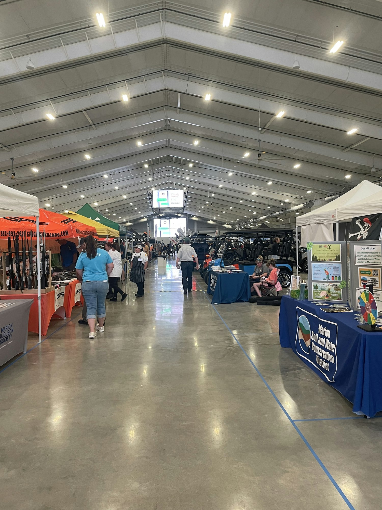 An indoor event with booths and visitors, featuring banners and a concrete floor, under a well-lit ceiling with hanging lights.