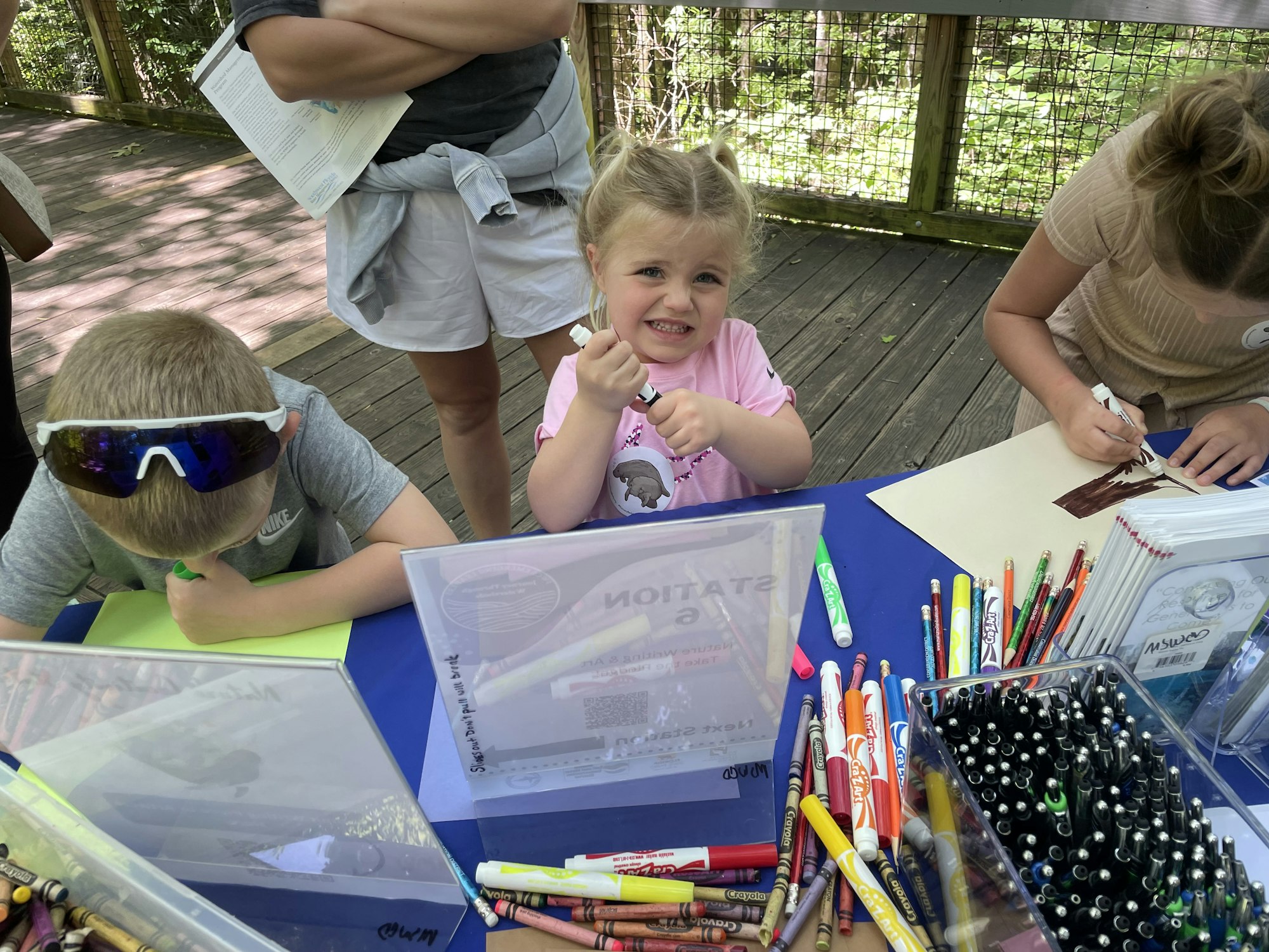 Kids drawing at a table with crayons and markers, outdoors on a sunny day.