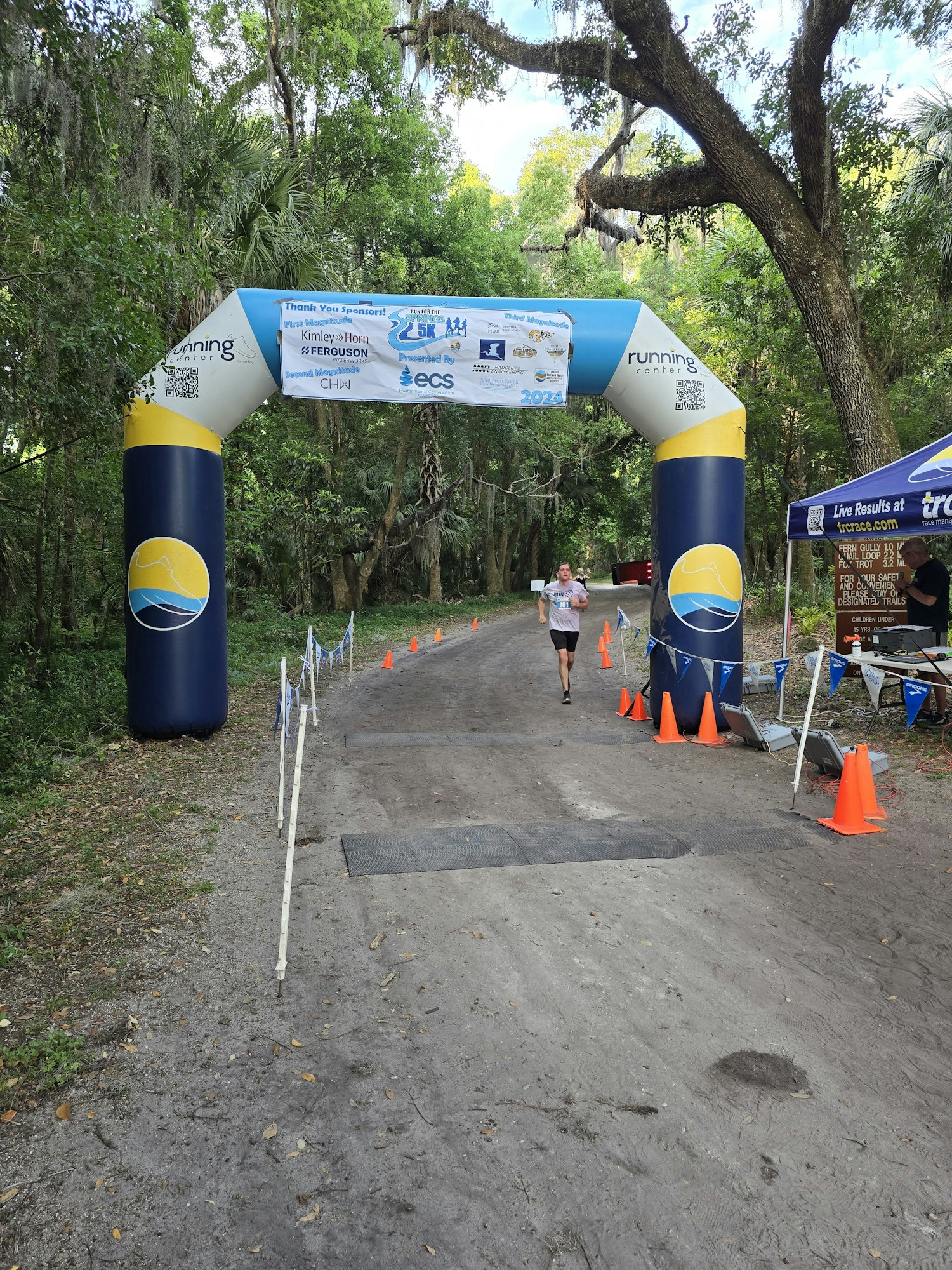 A person running through a race finish line arch in a park with trees and traffic cones. (Please note that the image is rotated 90 degrees.)
