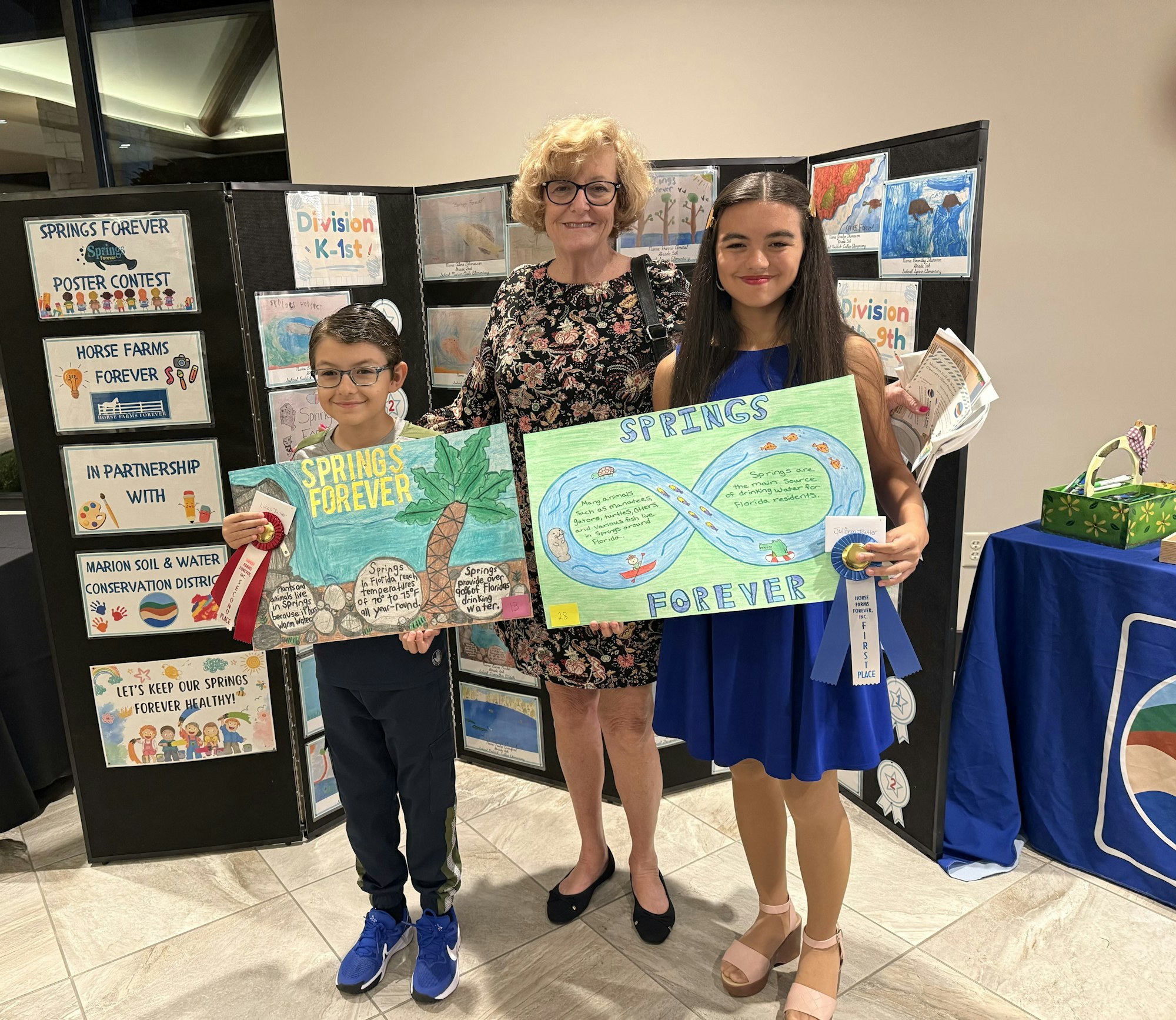 An award ceremony with two kids holding their winning posters and an adult, surrounded by conservation-themed displays.