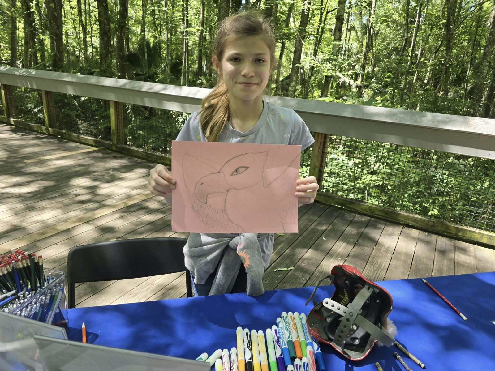 A young person is holding up a drawing of a cat's face, sitting at a table with markers, outdoors surrounded by trees.