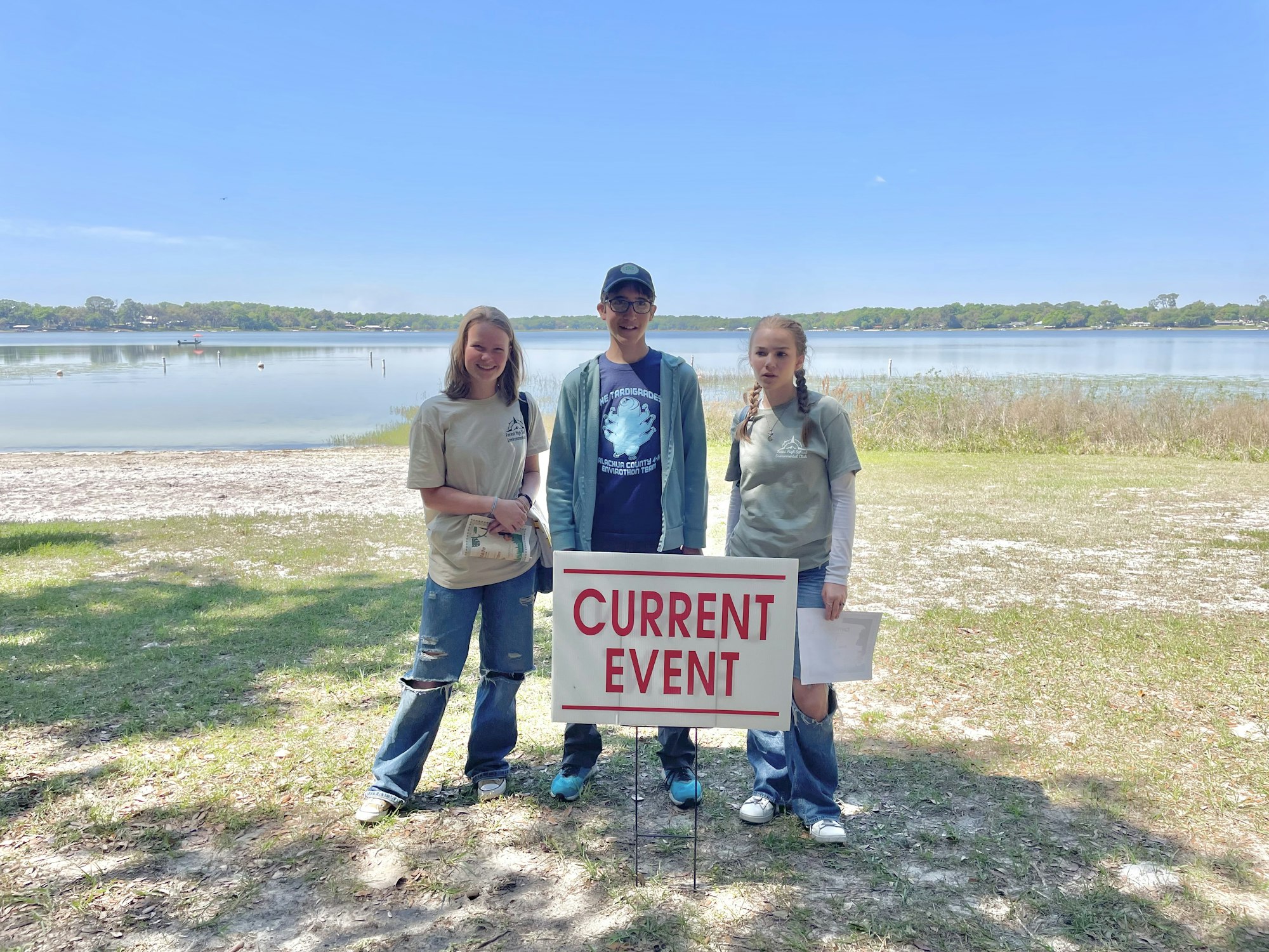 Three people stand by a "CURRENT EVENT" sign near a body of water.