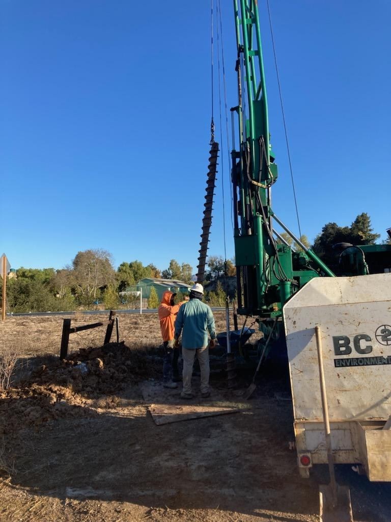 Workers operating a drilling rig with a large auger bit on a construction site under a clear blue sky.