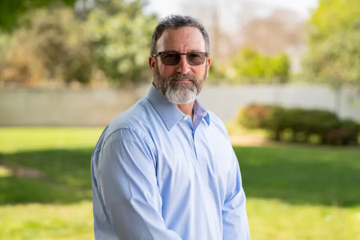 A man with a beard wearing glasses and a light blue shirt stands outdoors with greenery in the background.